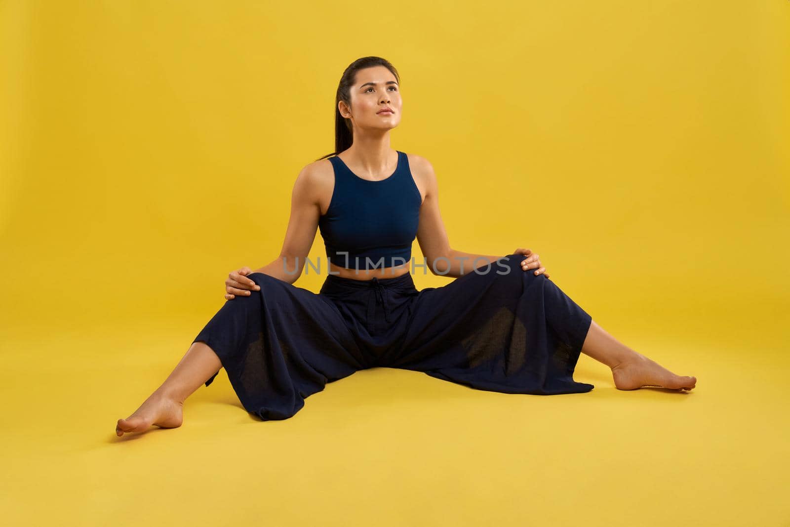 Young female yogi doing exercise, stretching legs in spacious studio. Slim caucasian woman with ponytail looking up, holding arms on apart knees, isolated on yellow studio background. Concept of yoga.