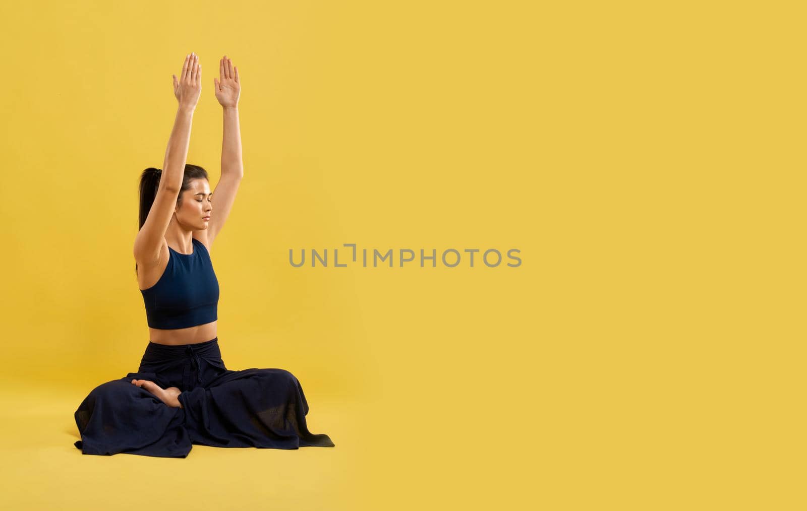 Calm girl meditating, with closed eyes alone on floor. Front view of female in black top sitting in lotus pose with gazed up hands, isolated on orange studio background, copy space. Concept of yoga.