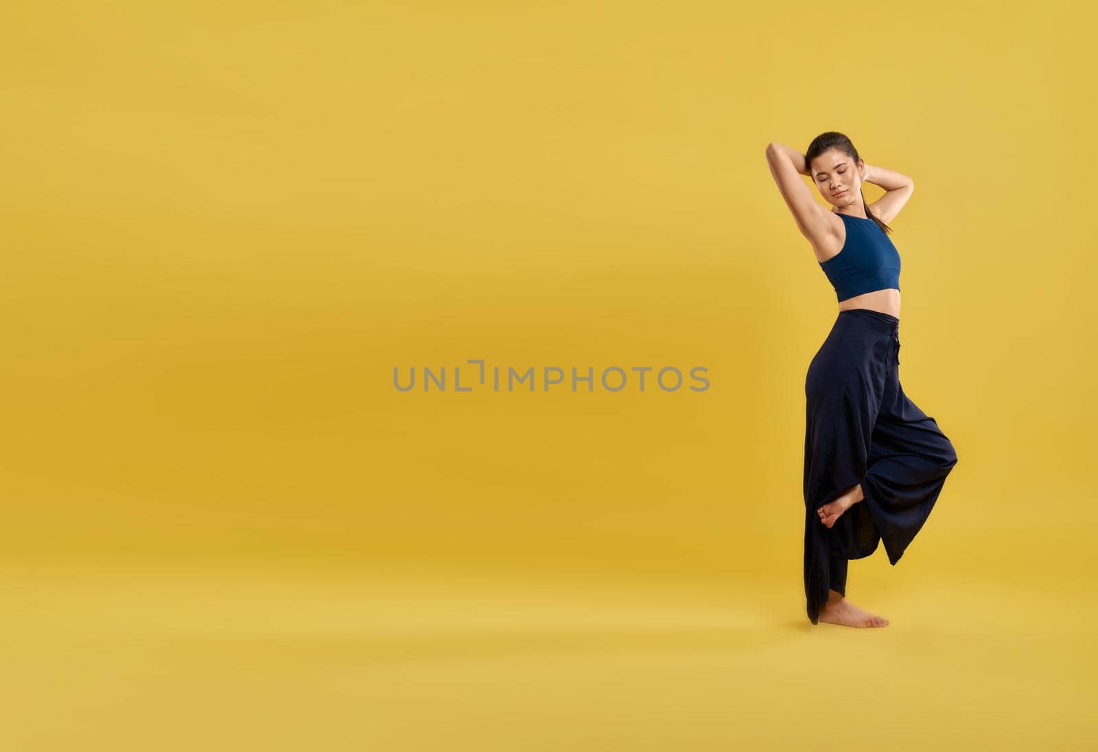 Female dancer with closed eyes keeping hands behind head while having rest indoors. Side view of cute girl smiling, while standing on one leg, isolated on yellow background. Concept of relaxation.