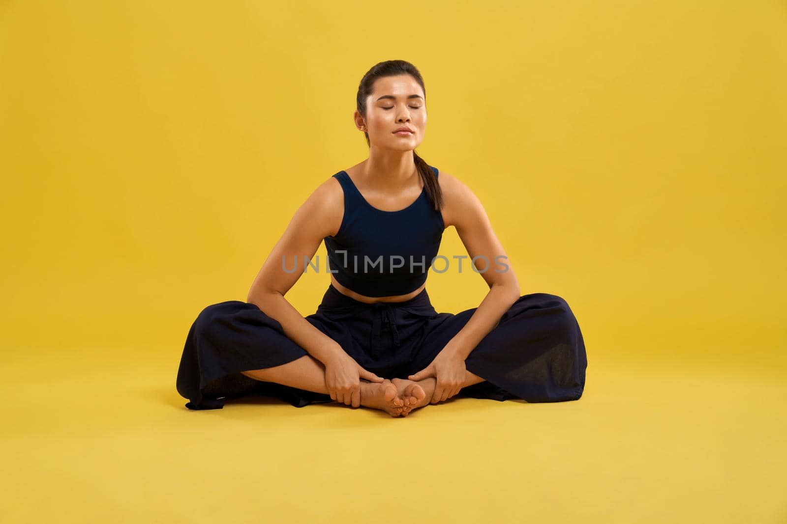 Pretty girl meditating in butterfly II yoga pose, with closed eyes while exercising in studio. Front view of barefoot lady practicing yoga, isolated on orange studio background. Concept of yoga asana.