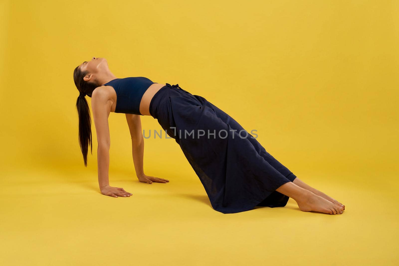 Fit caucasian girl stretching body at upward plank pose during training in studio. Side view of brunette woman in sportswear practicing yoga, isolated on orange studio background. Concept of yoga.