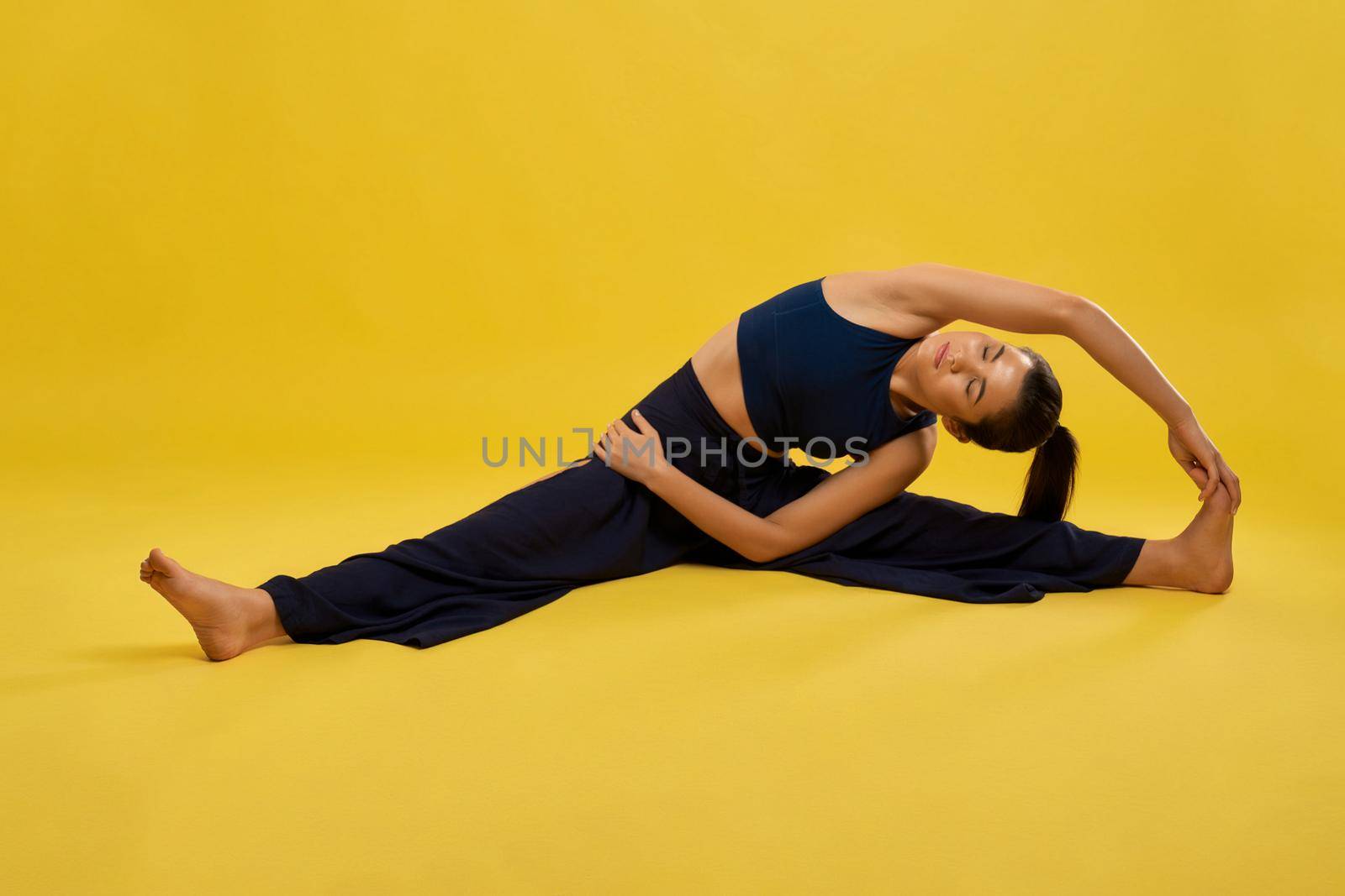 Pretty woman practicing yoga asana with stretching, leaning to leg indoors. Front view of barefoot girl with closed eyes doing twine, isolated on yellow studio background. Concept of yoga exercising.