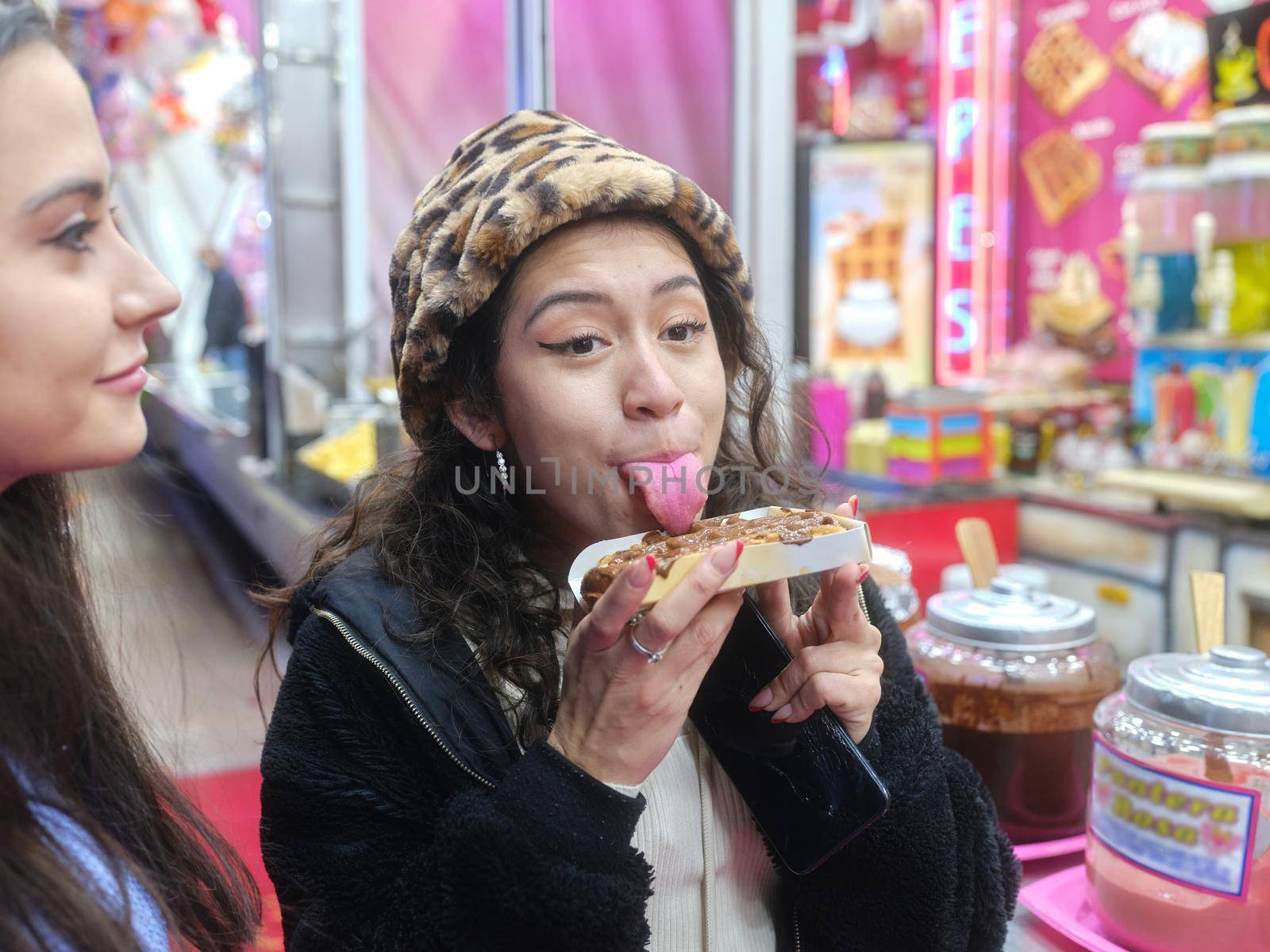 Latina woman in leopard hat licking a cocholate waffle while standing in a night fair with a friend