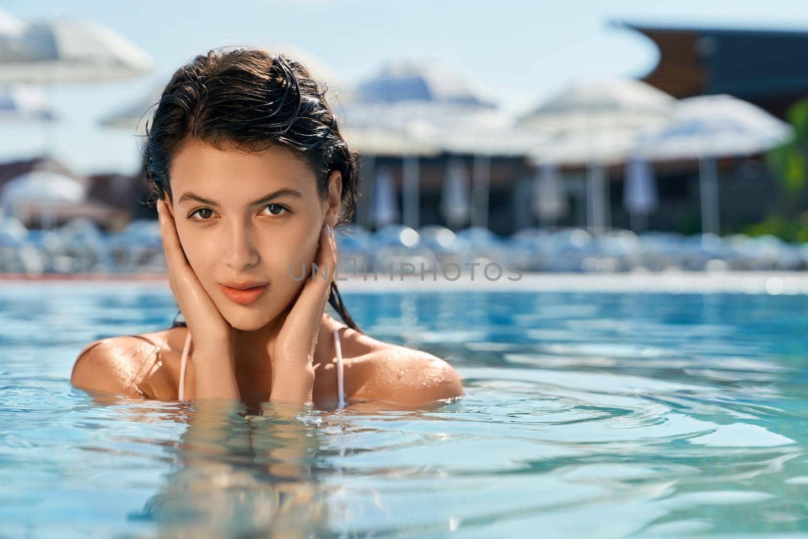 Front view of pretty young female with long hair standing in water. Brunette girl wearing white swimsuit swimming in pool, touching hair, looking at camera. Concept of youth.