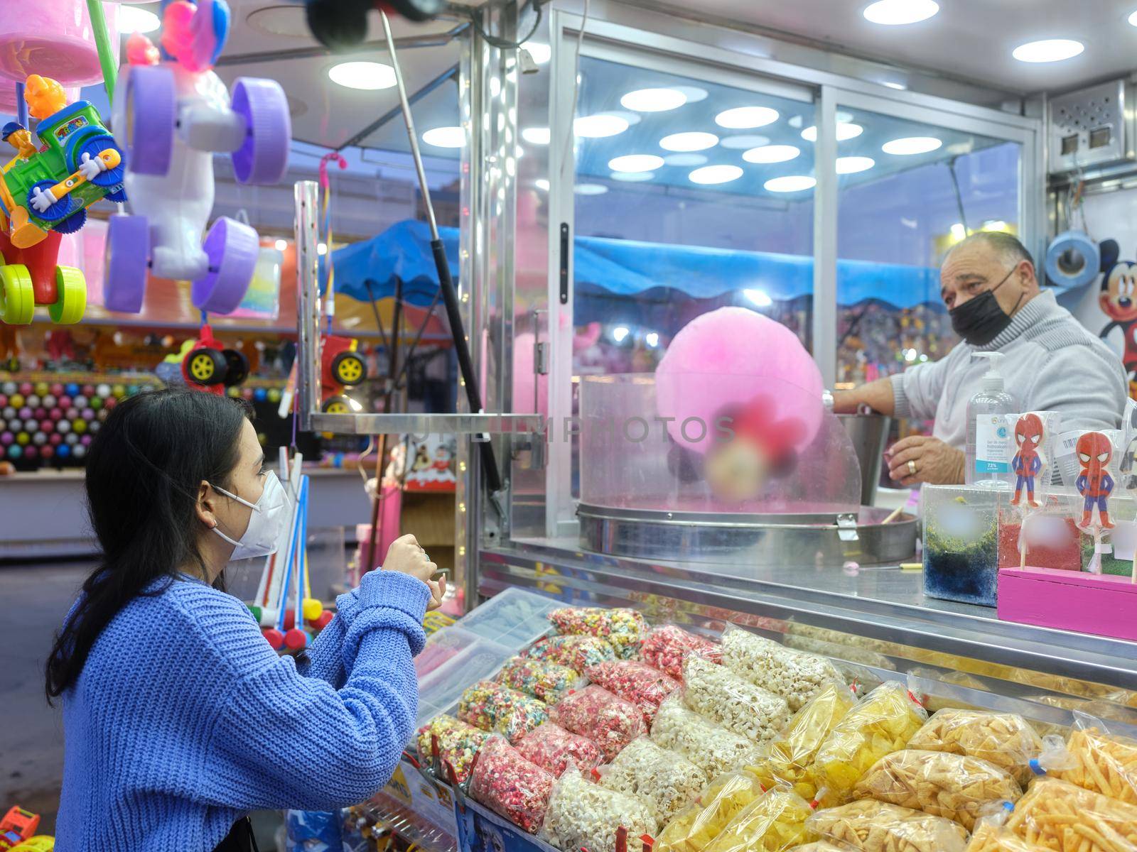 Young latin woman in mask buying cotton candy in an evening fair