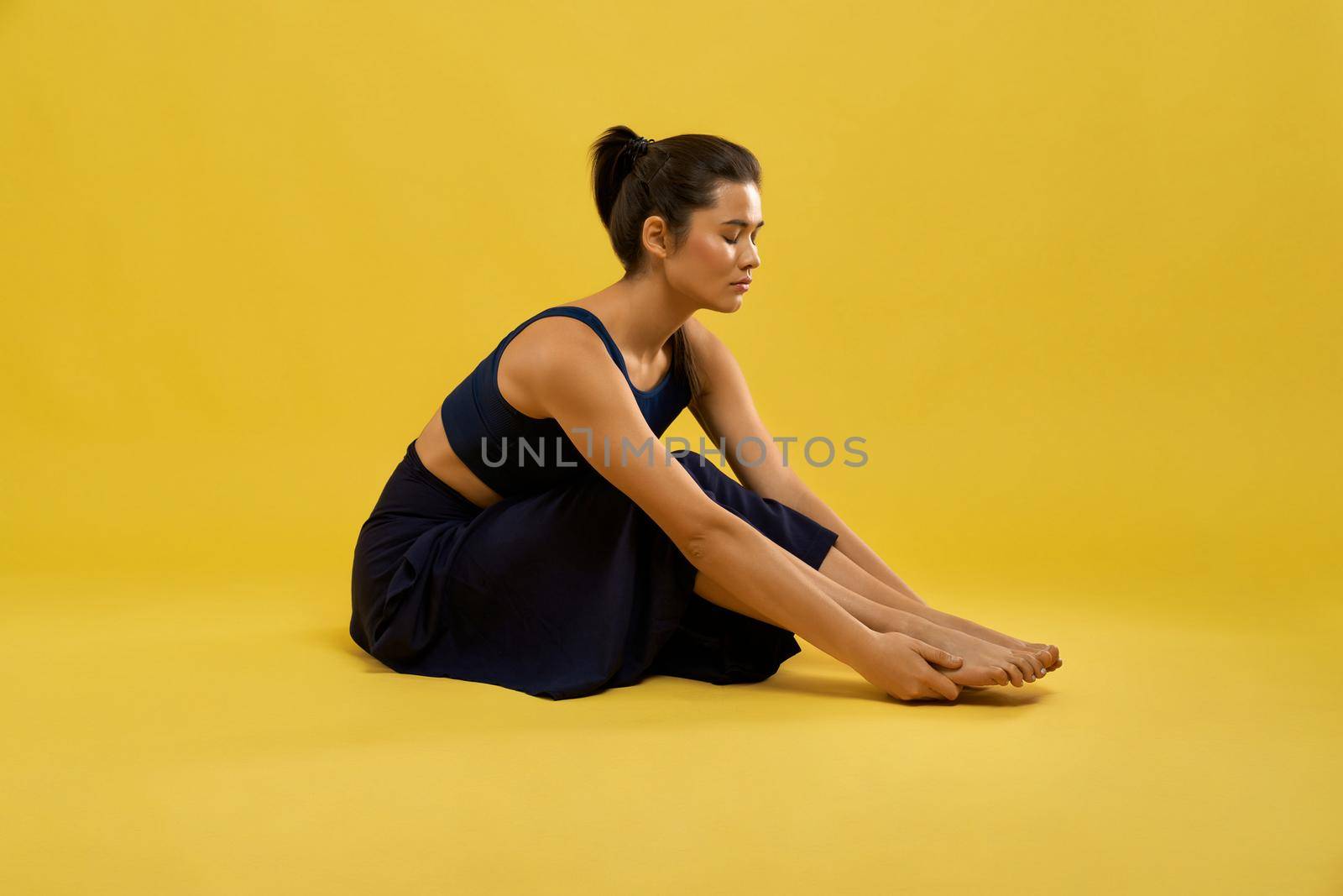 Young woman holding her feet while doing stretching exercises in studio. Side view of serious girl sitting in forward bend, keeping feet with hands, isolated on orange background. Concept of exercise.