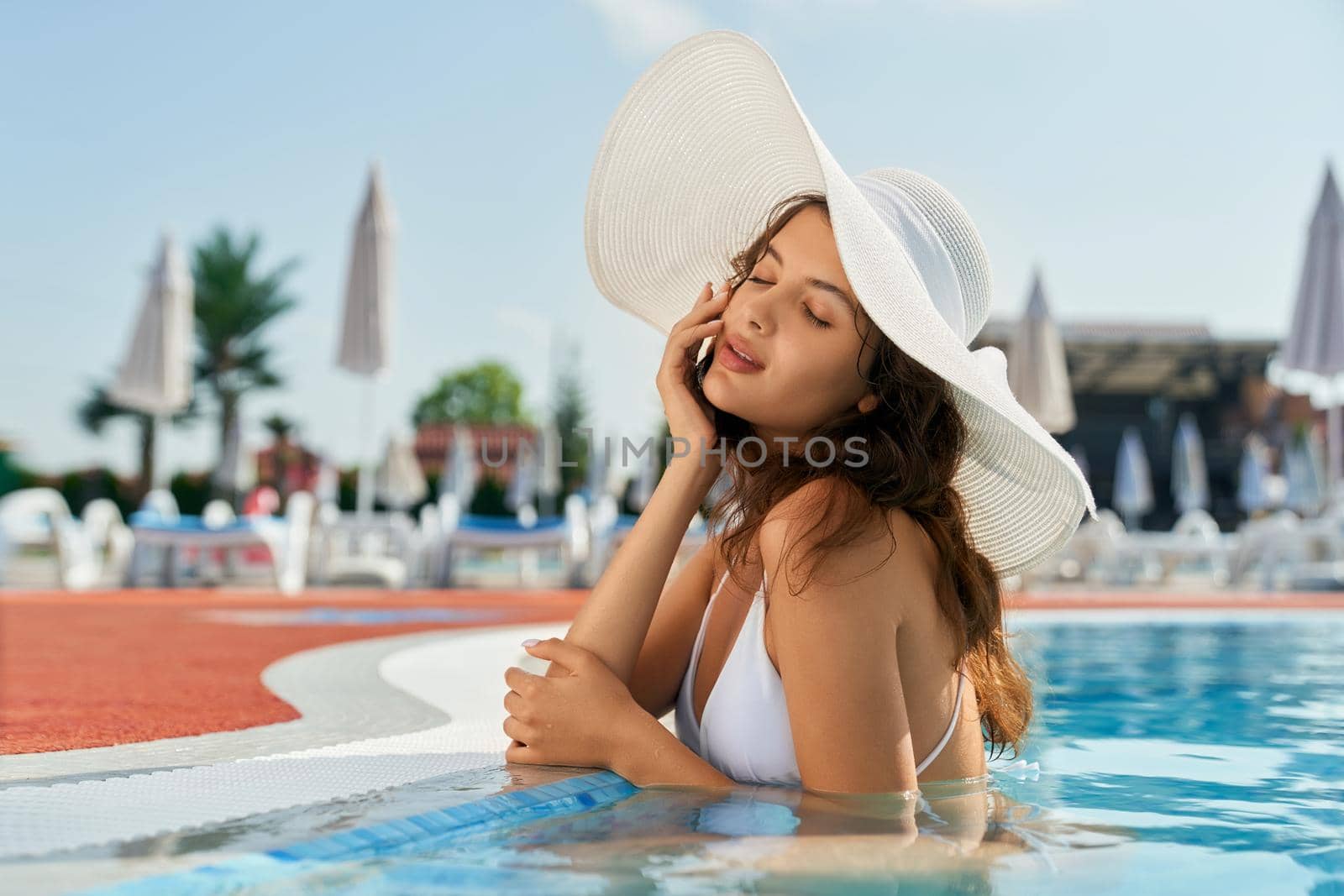 Side view of pretty girl wearing white swimsuit and hat standing in pool. Young female swimming, relaxing, sunbathing with closed eyes, dreaming. Concept of summertime and youth.