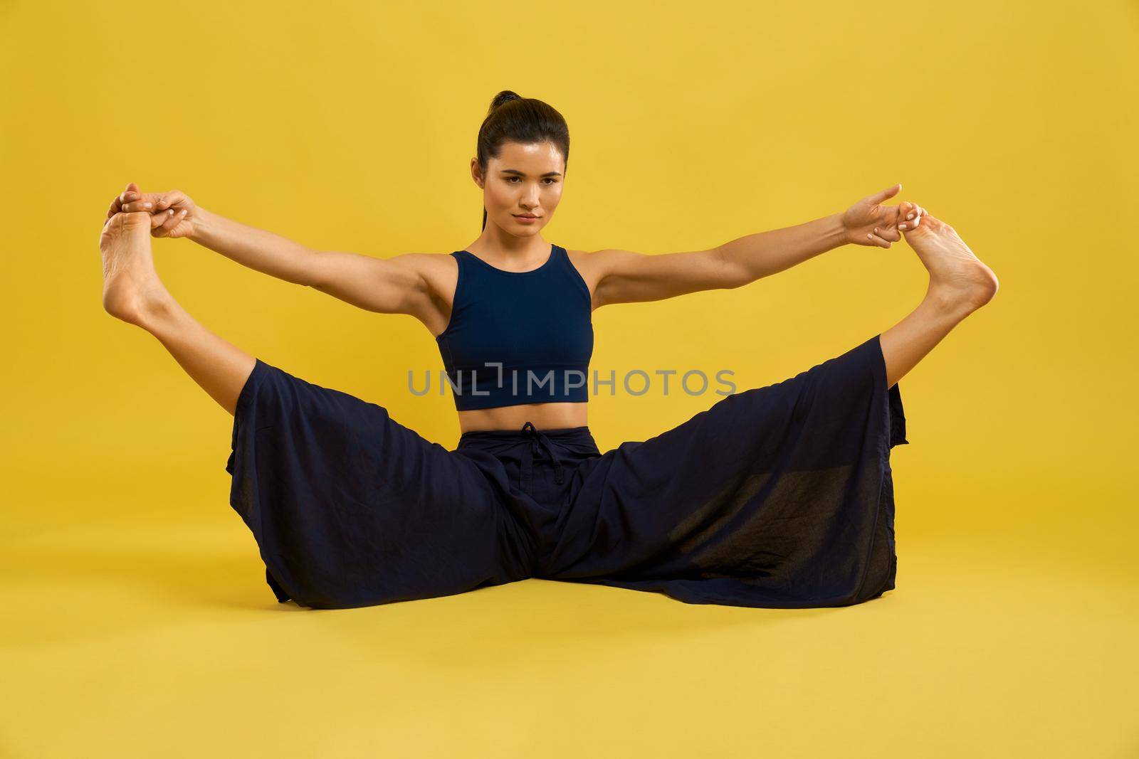 Fit caucasian girl stretching legs in Upavistha Konasana during yoga training. Front view of brunette woman in wide pants practicing yoga, isolated on tangerine studio background. Concept of yoga.