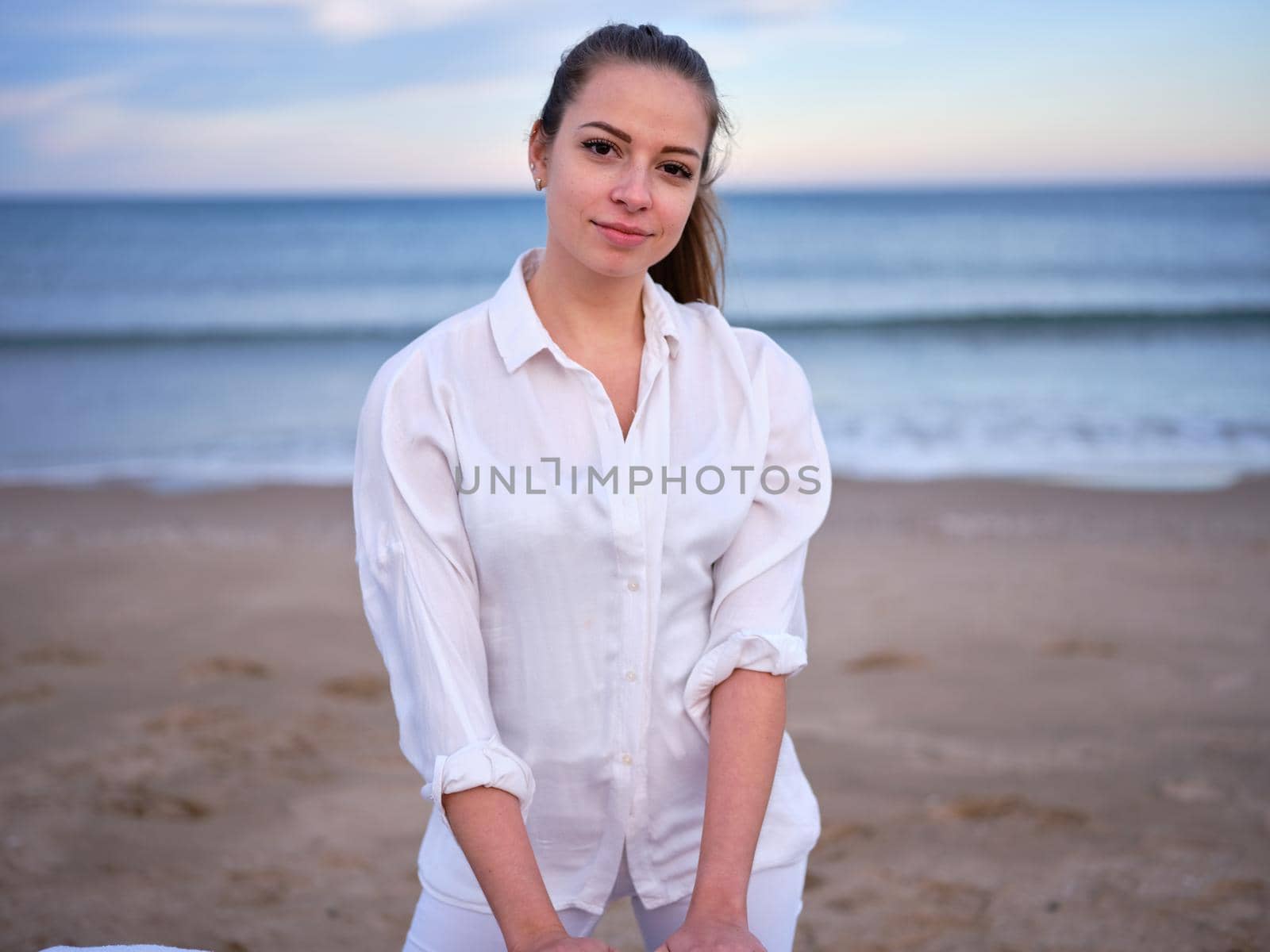 Front view of a young chiromassage therapist dressed in white standing on a beach in Valencia with the sea in the background.