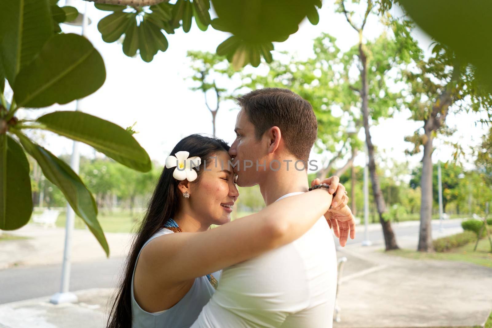 South african man kissing the forehead of a thai woman with a flower in the hair in a park