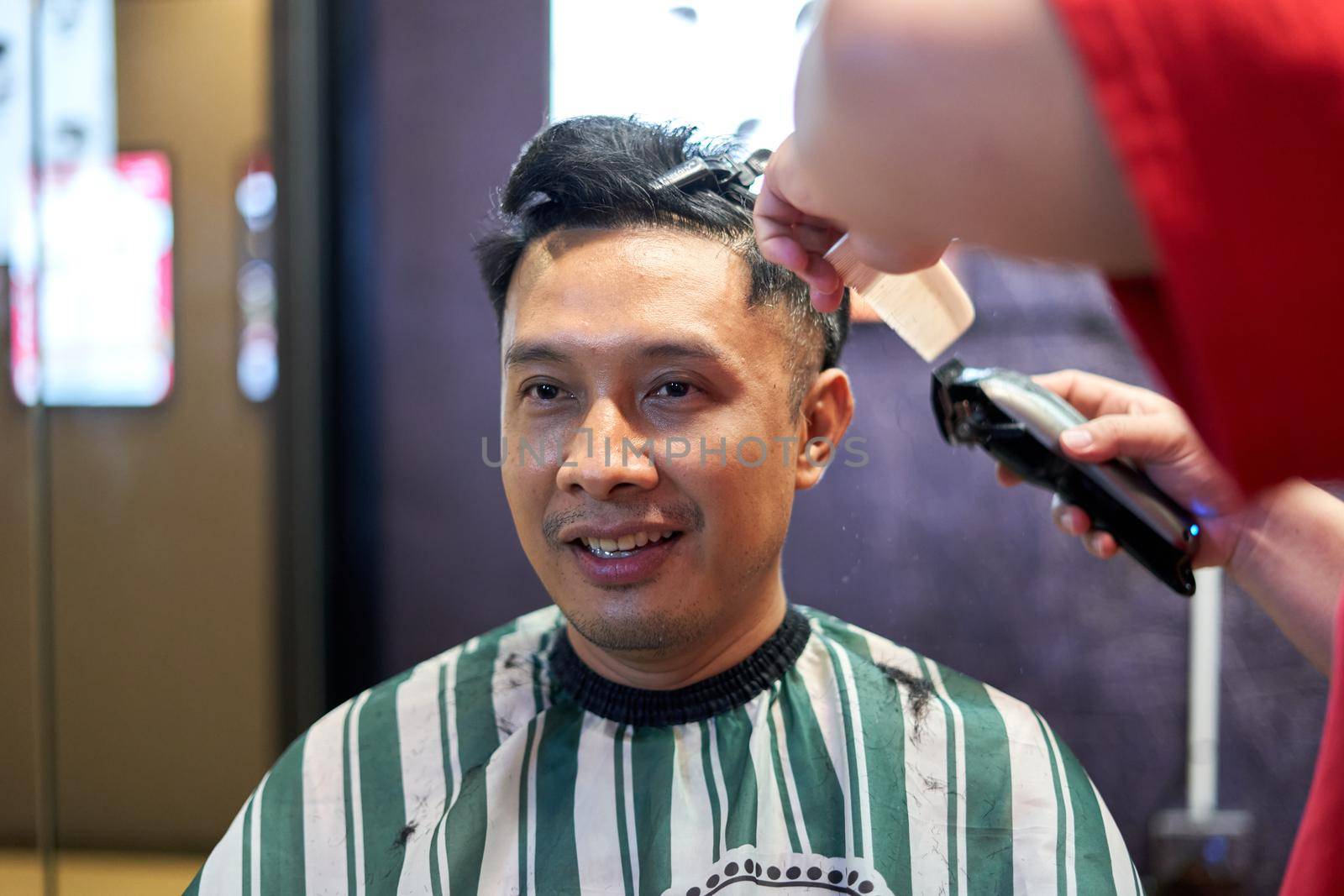 Portrait of a smiling asian man sitting on a barber shop while the barber cutting his hair