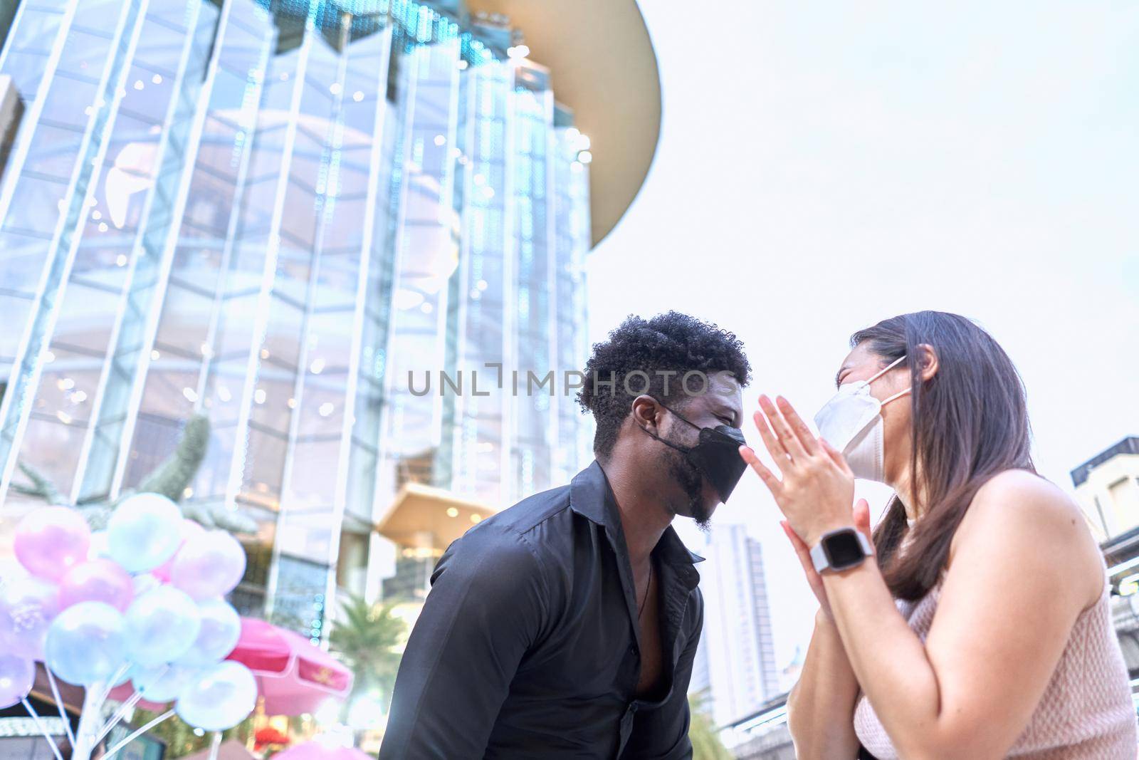 Two multiethnic friends in mask laughing outside a shopping mall with a skyscraper in the background