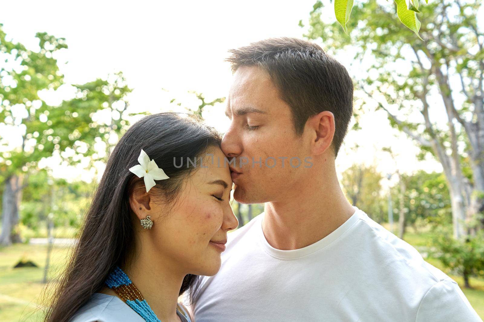 Close up view of a man kissing the forehead of a thai woman with a flower in the hair in a park