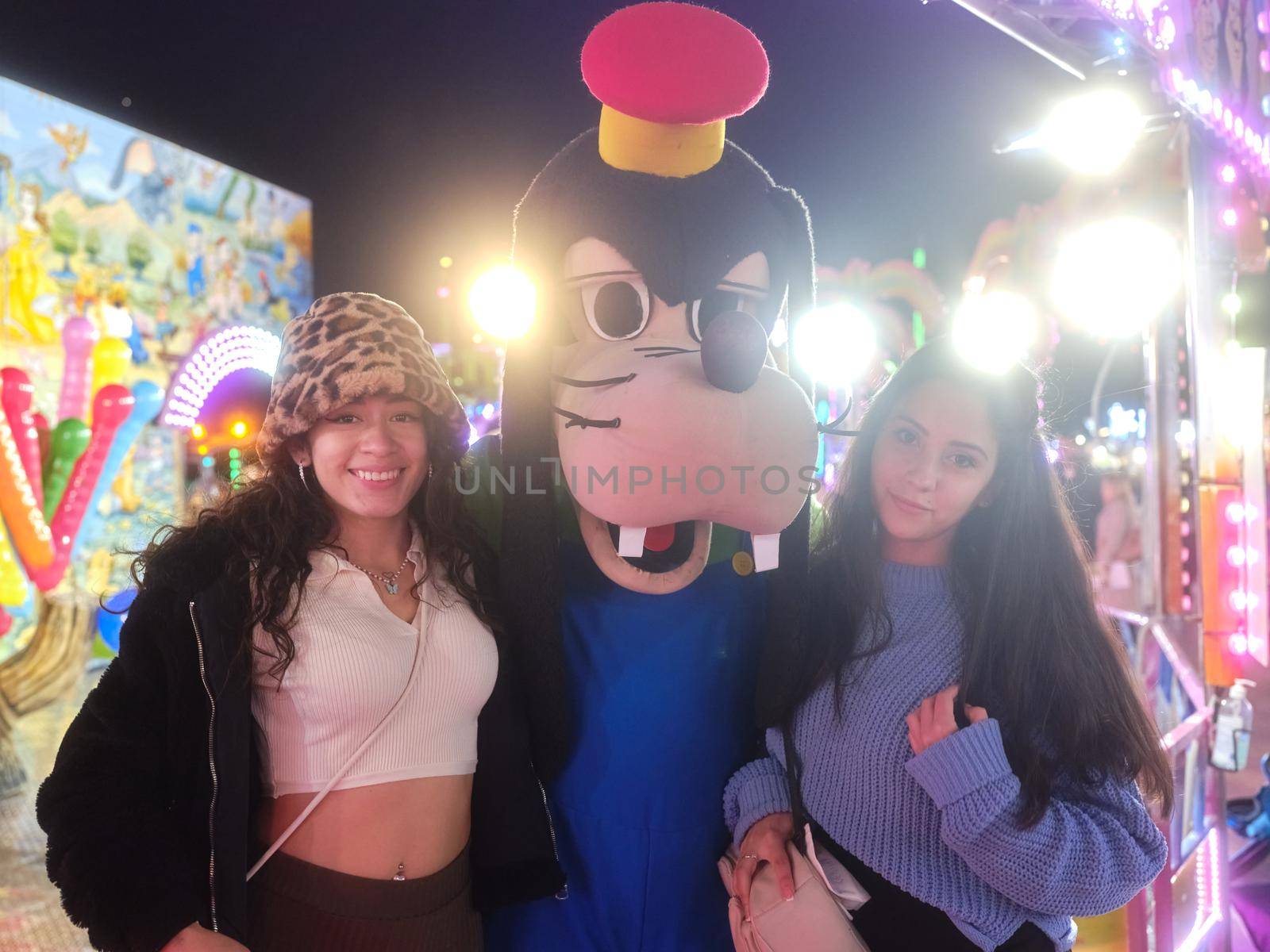 Portrait of two women standing next to a actor dressing like Goofy dog in a night fair