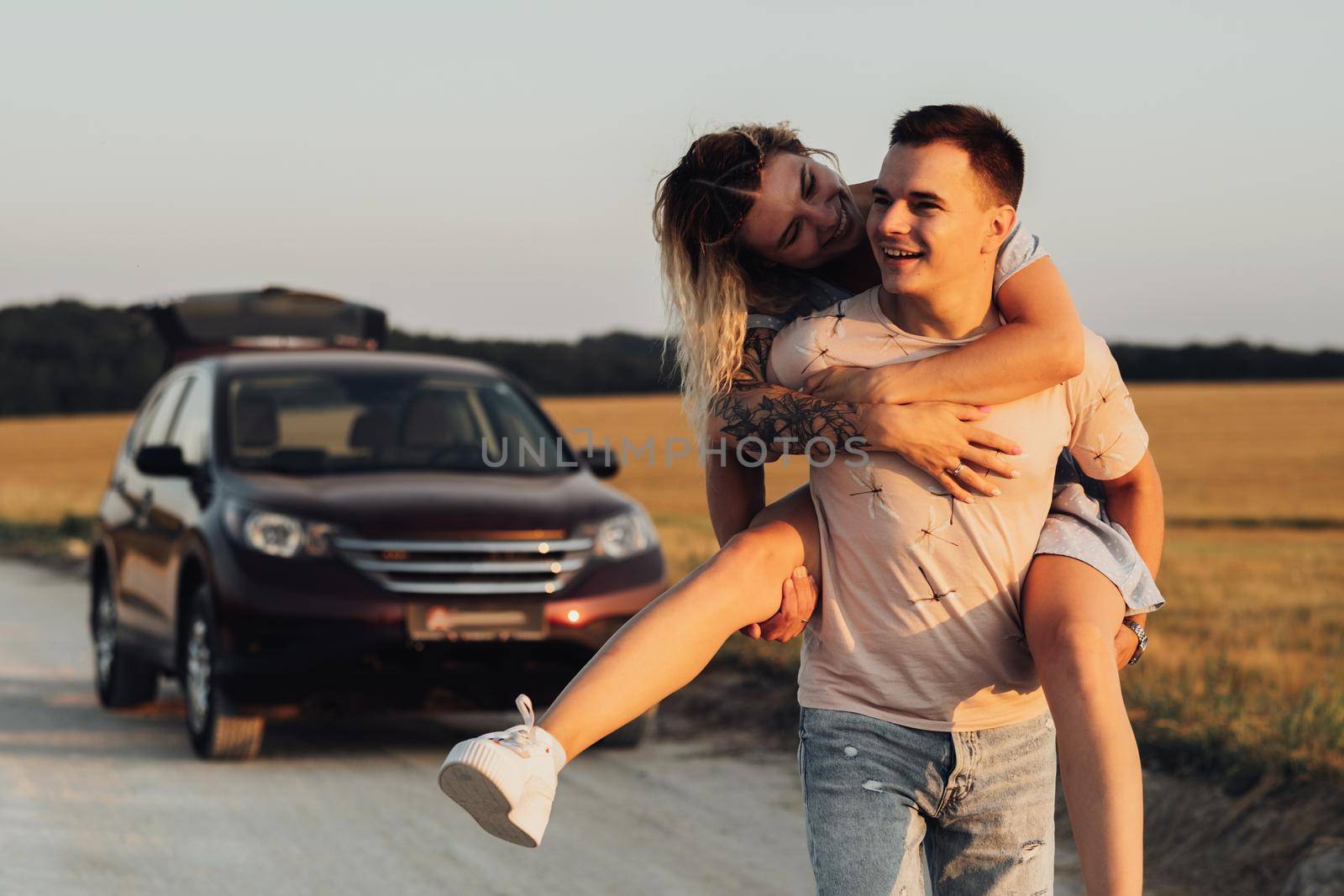 Caucasian Man Carries Girlfriend on His Back on Background of Their SUV Car, Young Couple Enjoying Their Road Trip at Sunset