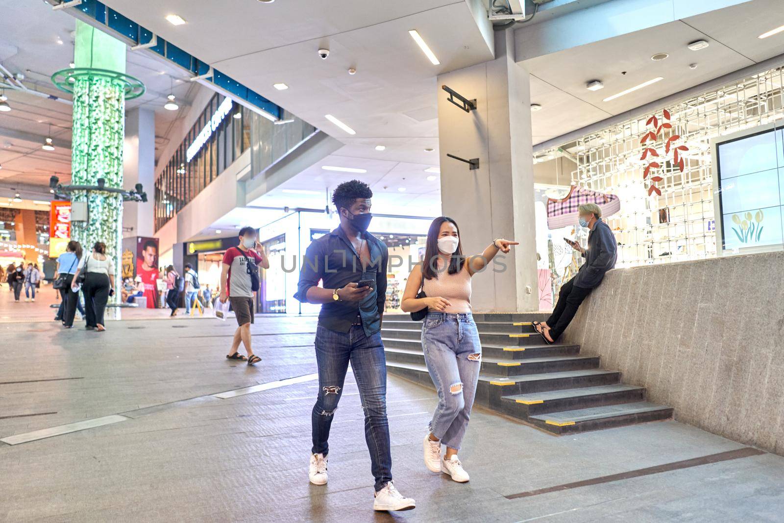 Asian woman wearing mask pointing ahead while walking with an african friend in a mall in Bangkok