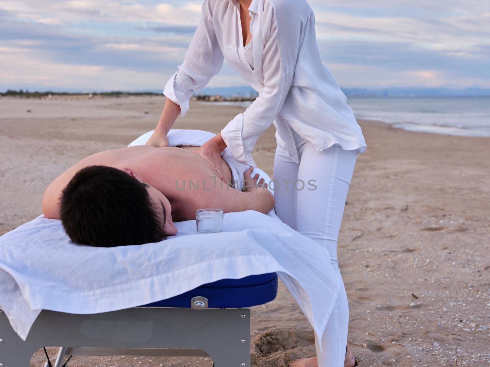 Front view of a young man lying on a massage table receiving a chiromassage treatment on the beach in Valencia by an unrecognizable young chiromassage therapist.