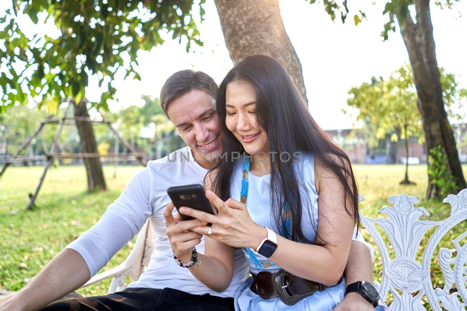 Smiling multiracial newlywed couple using the mobile while sitting on a bench outdoors during sunset