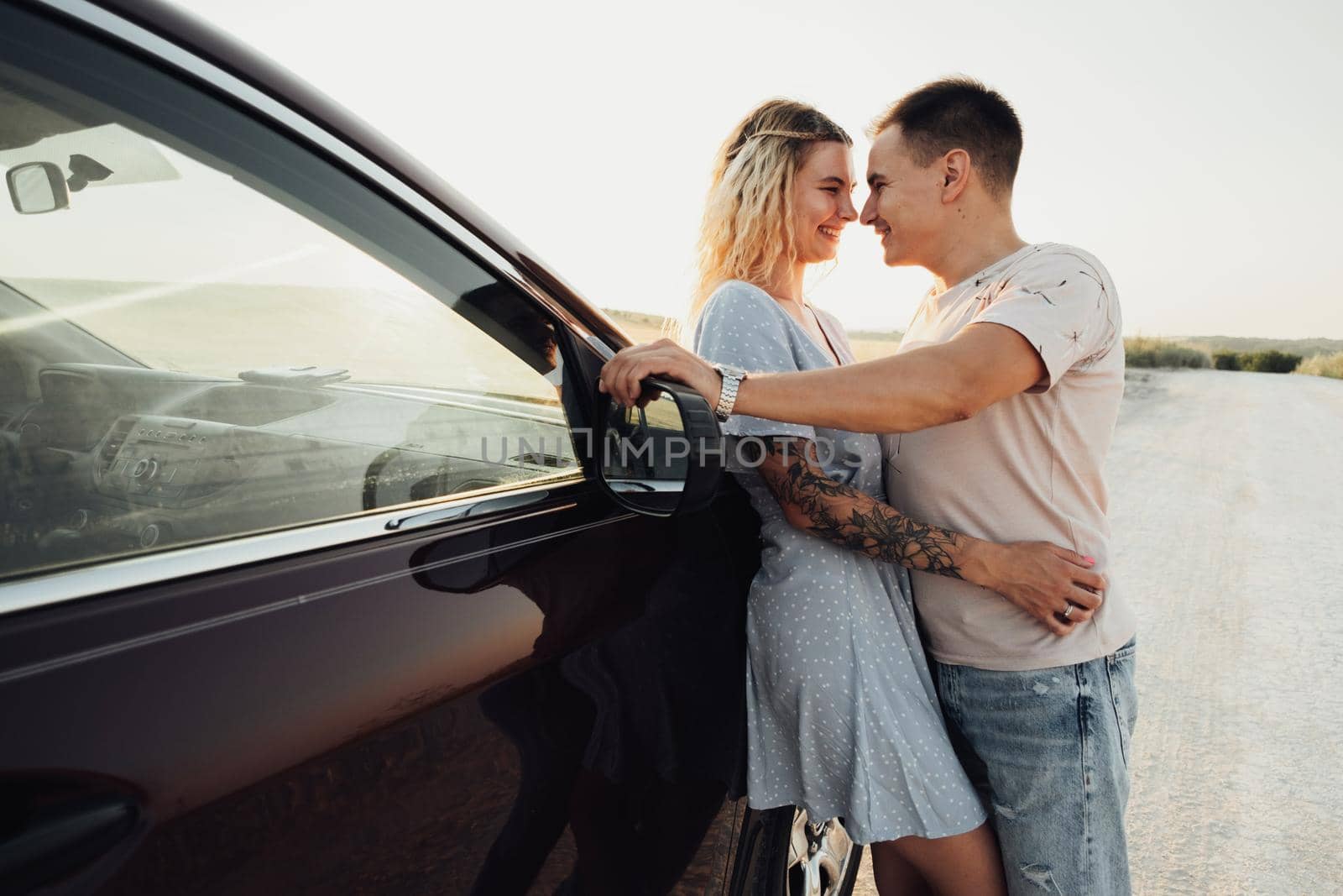 Cheerful Young Couple Hugging Near the Car, Tattooed Woman and Caucasian Man Enjoying Road Trip at Sunset by Romvy