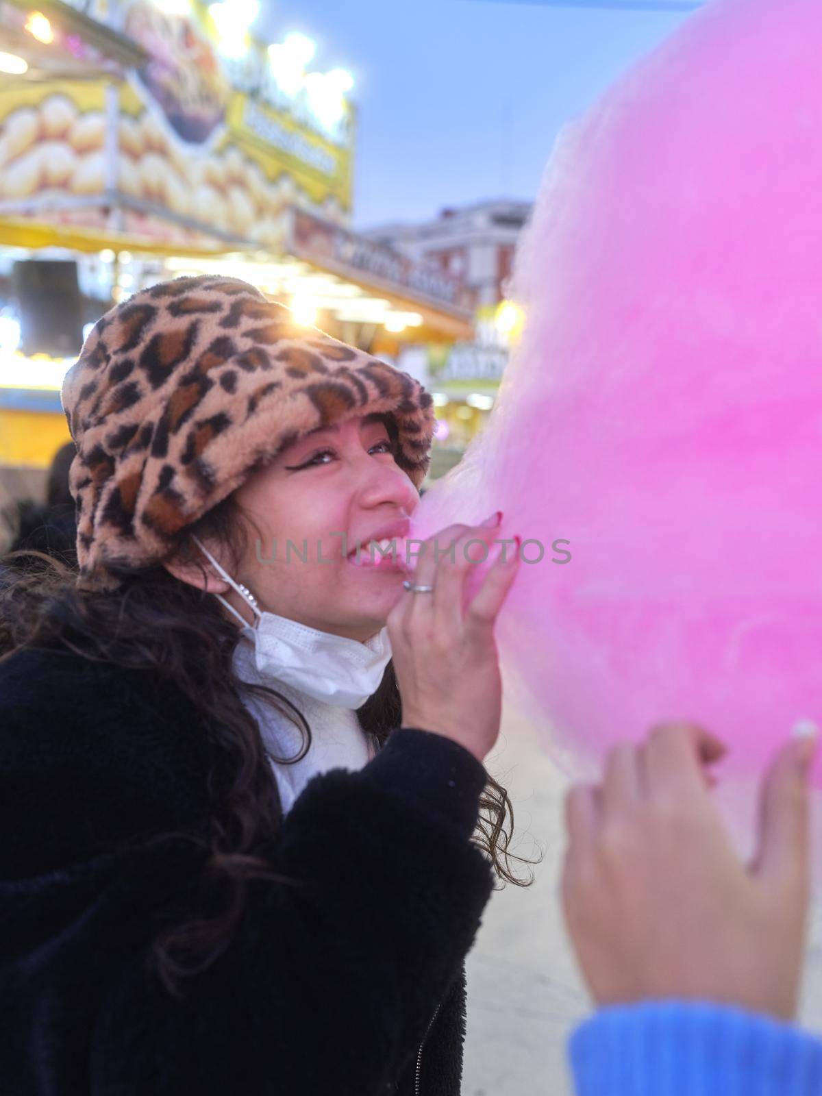 Vertical photo of a latina young woman biting a piece of pink cotton candy during a night fair