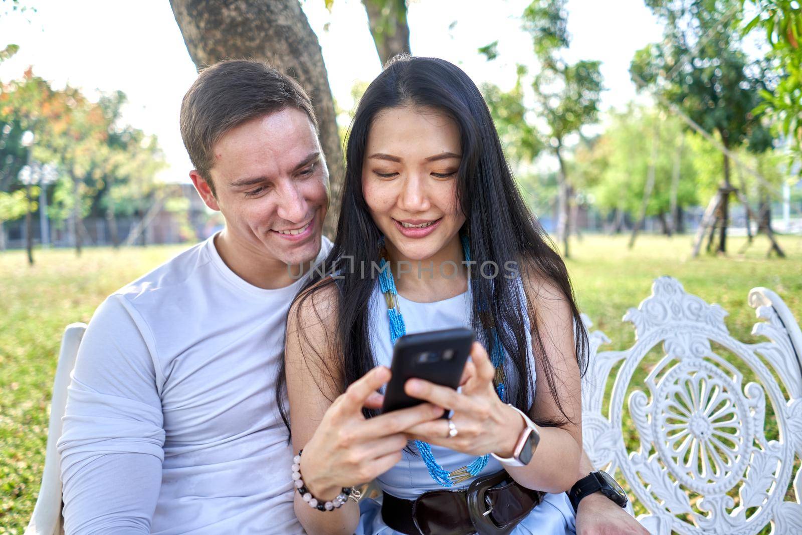 Asian woman showing the screen of the mobile to a man sitting together on a bench in a park