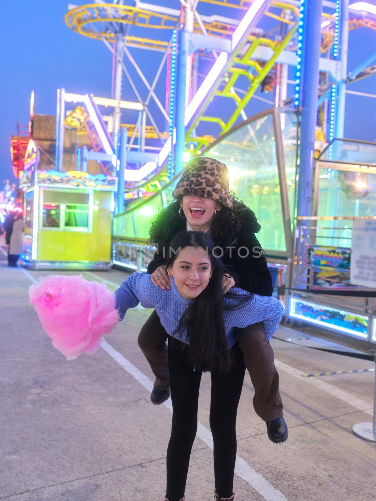 Female friend carrying other in the back in the middle of a night fair while holding a cotton candy