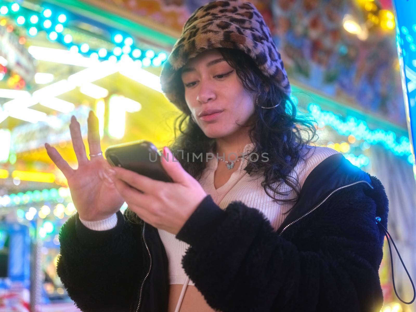 Latina woman in hat typing on her mobile phone while standing in front of a fairground ride at night