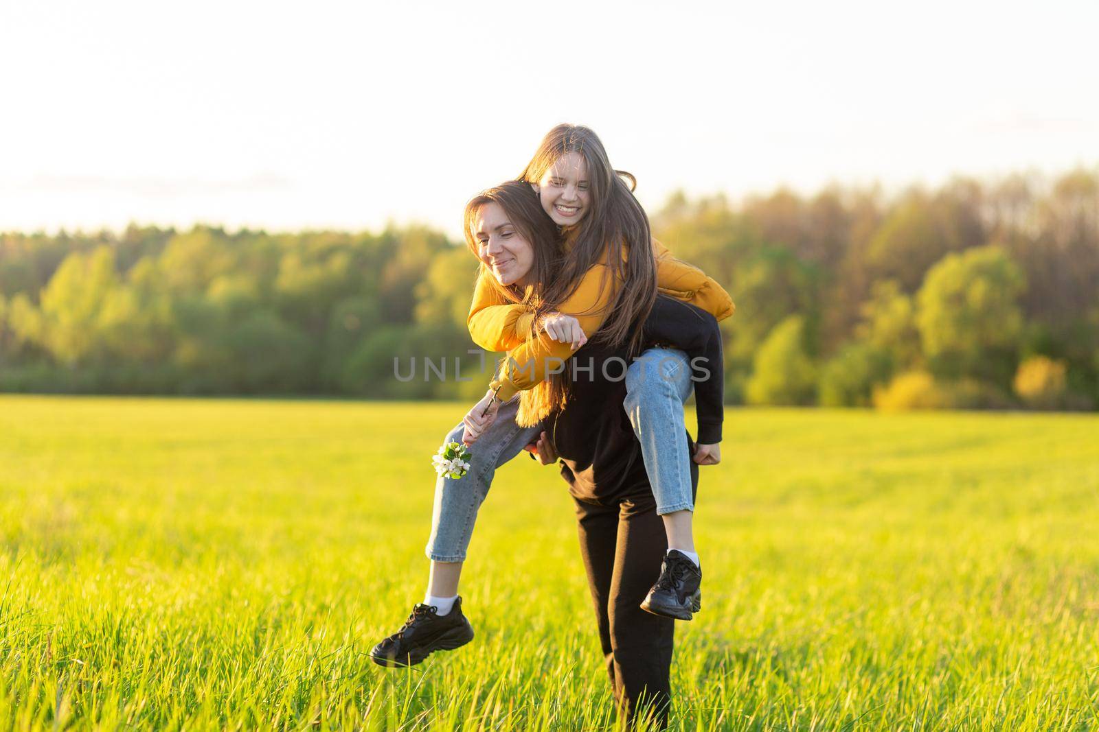 Playful mother giving daughter piggy back ride at green field. Both laughing and look happy. Spring in forest background. Closeup.