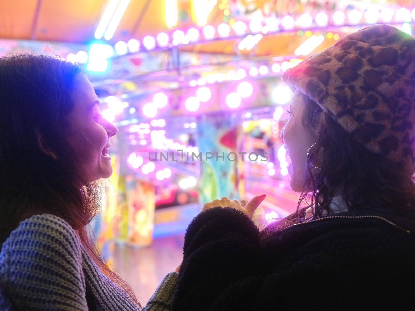 Two women talking in front of an attraction of a colorful evening fair by WesternExoticStockers
