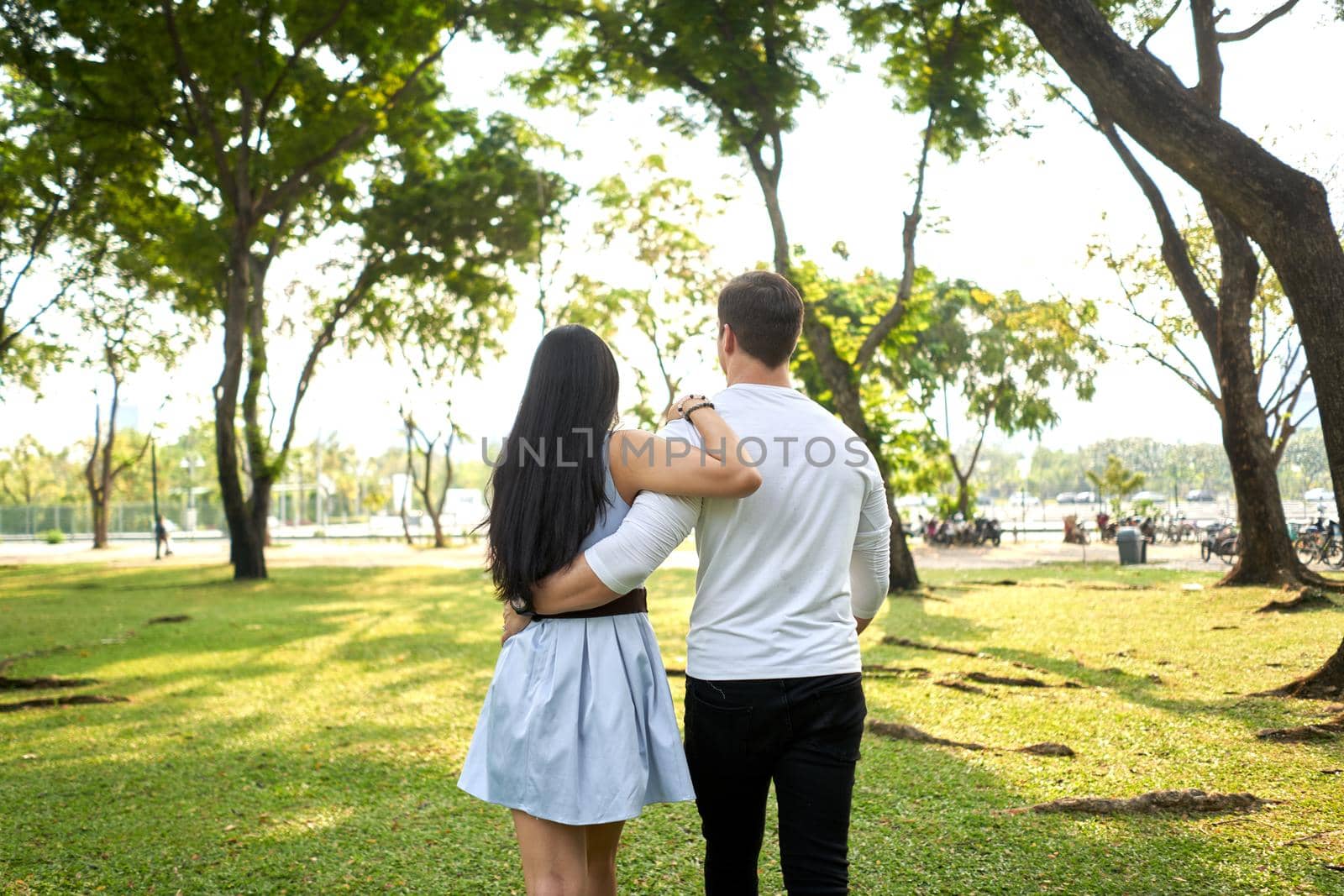 Back of multiracial couple walking in an urban park holding each other's backs by WesternExoticStockers