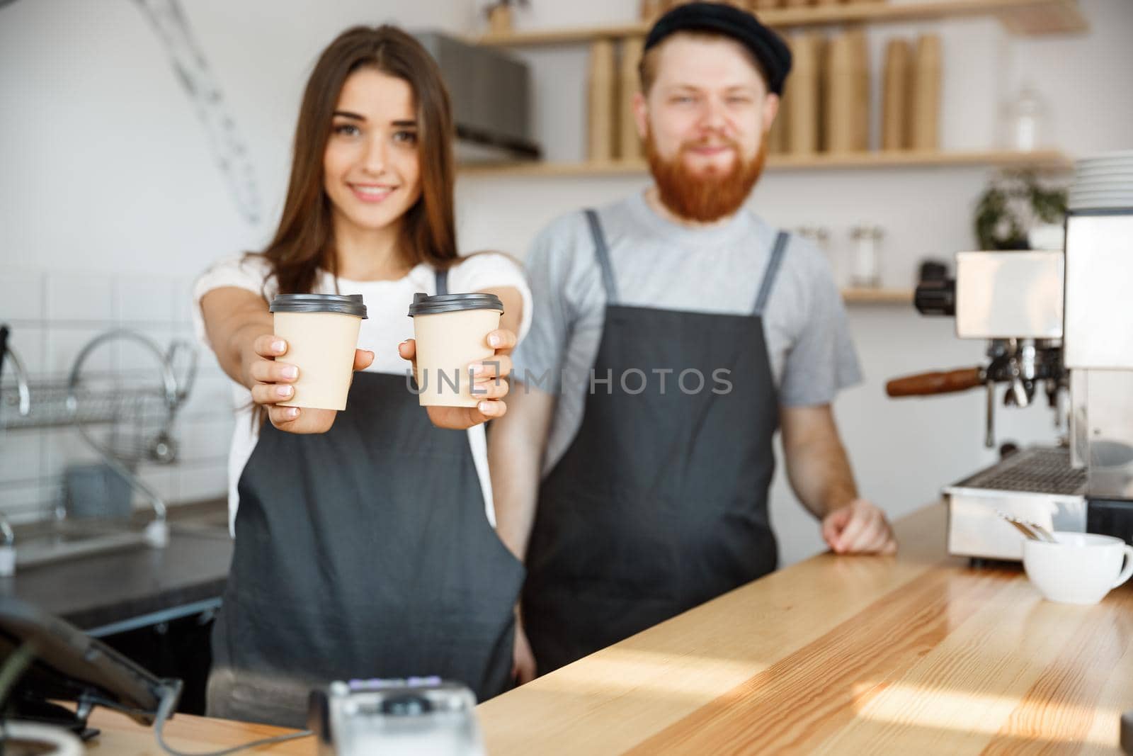 Coffee Business Concept - Positive young bearded man and beautiful attractive lady barista couple giving take away cup of coffee to custome at the modern coffee shop by Benzoix