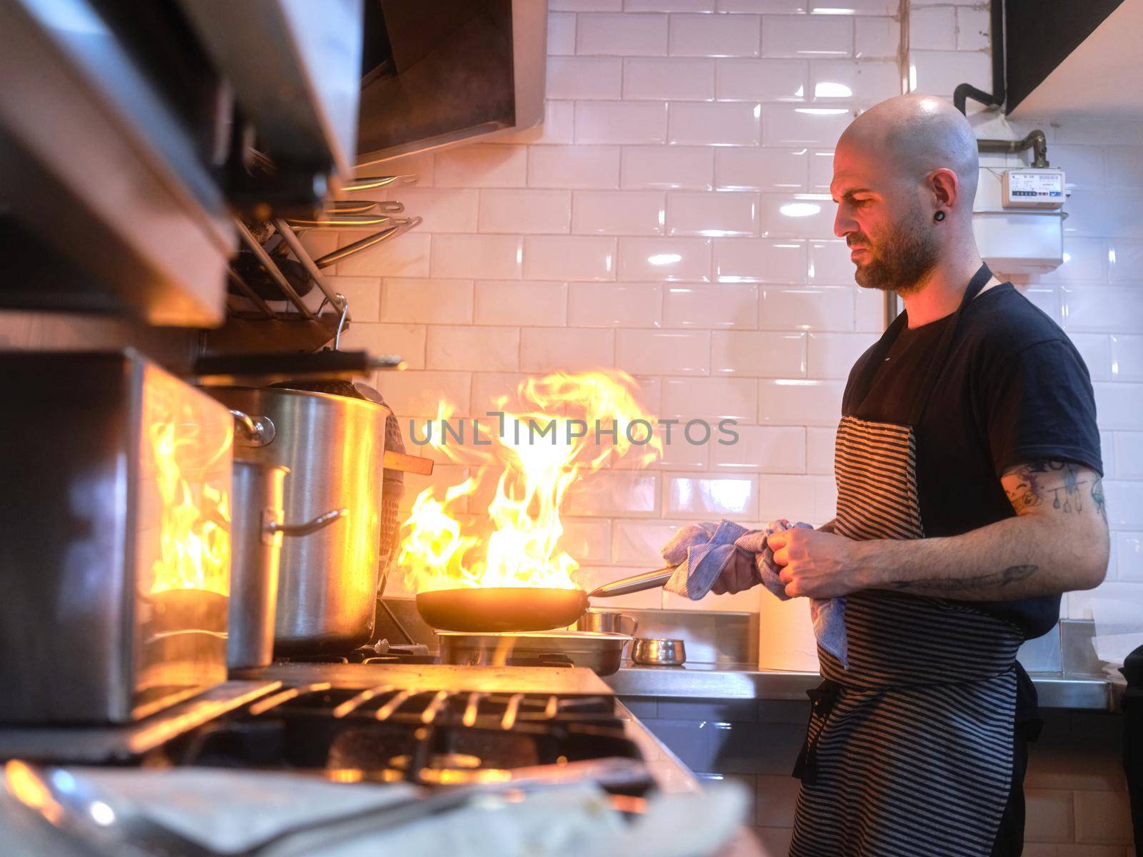 Chef in apron flaming the food in a saucepan in the kitchen of a restaurant by WesternExoticStockers