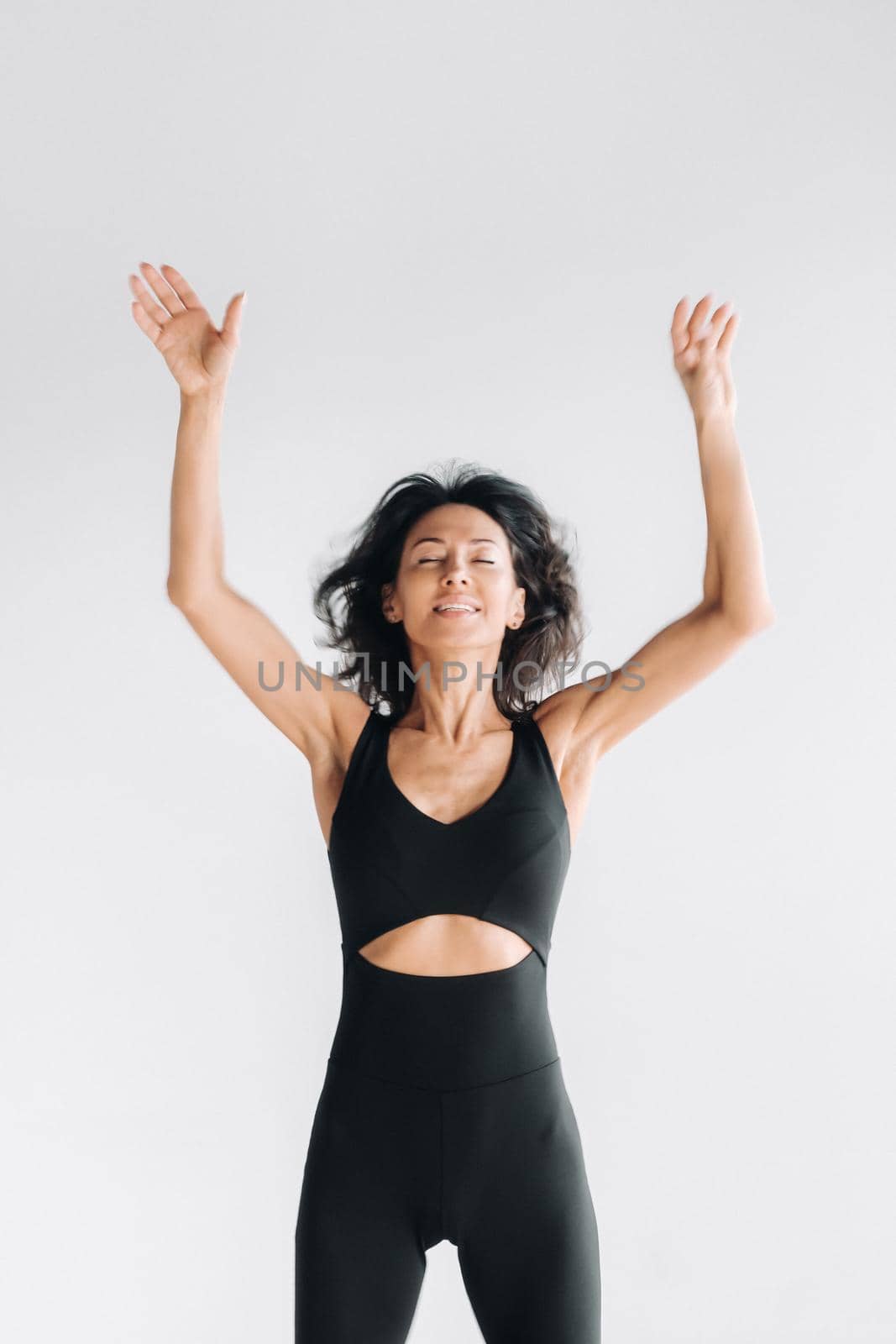 A blurry silhouette of a woman in black sportswear jumps up and raises her arms up against a white background by Lobachad