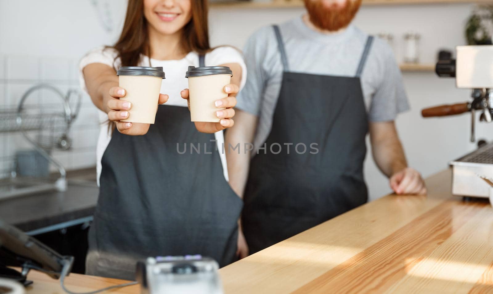 Coffee Business Concept - Positive young bearded man and beautiful attractive lady barista couple giving take away cup of coffee to custome at the modern coffee shop