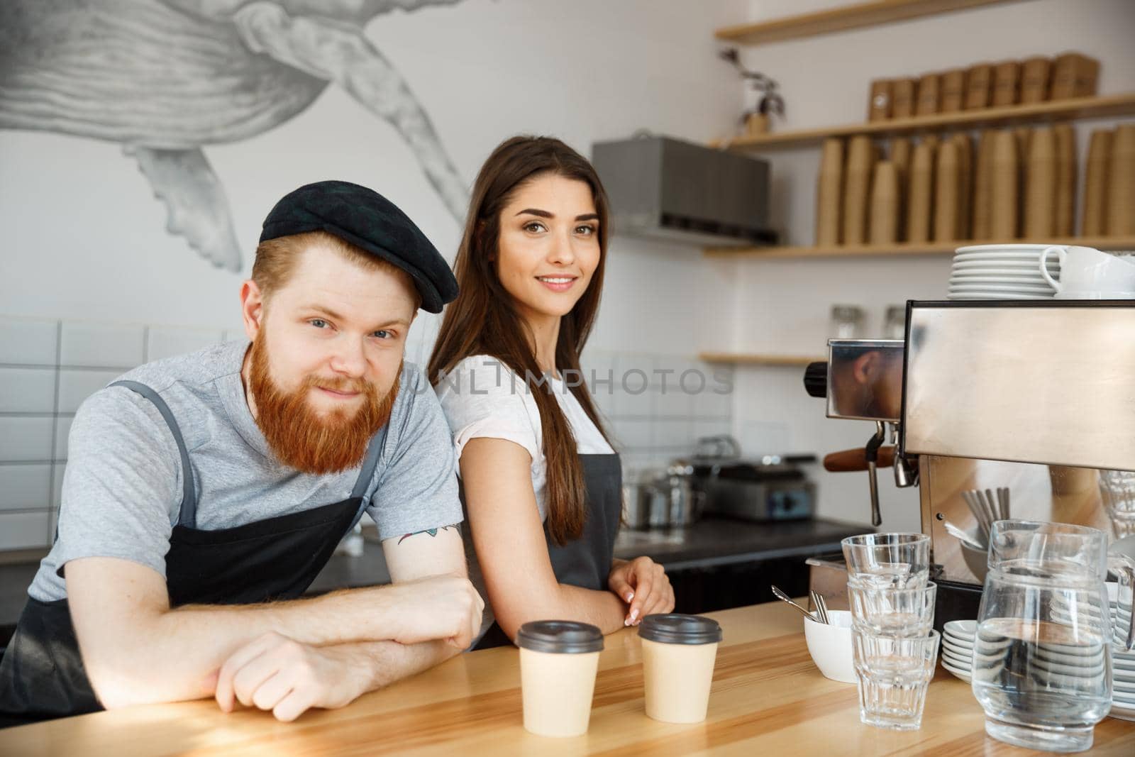 Coffee Business Concept - Positive young bearded man and beautiful attractive lady barista couple in apron while standing at bar Couter ready to give Coffee Service at the modern coffee shop by Benzoix