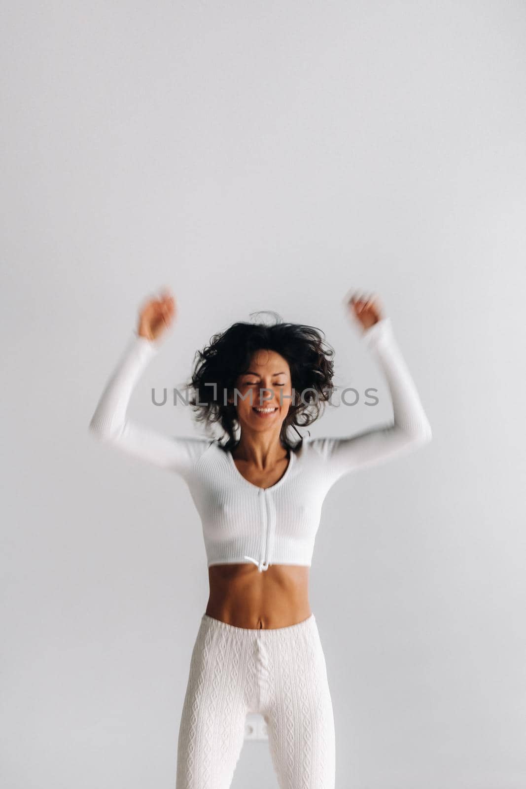 A blurry silhouette of a woman in white sportswear bounces on a white background. The girl, jumping, raised her hands up in the gym by Lobachad