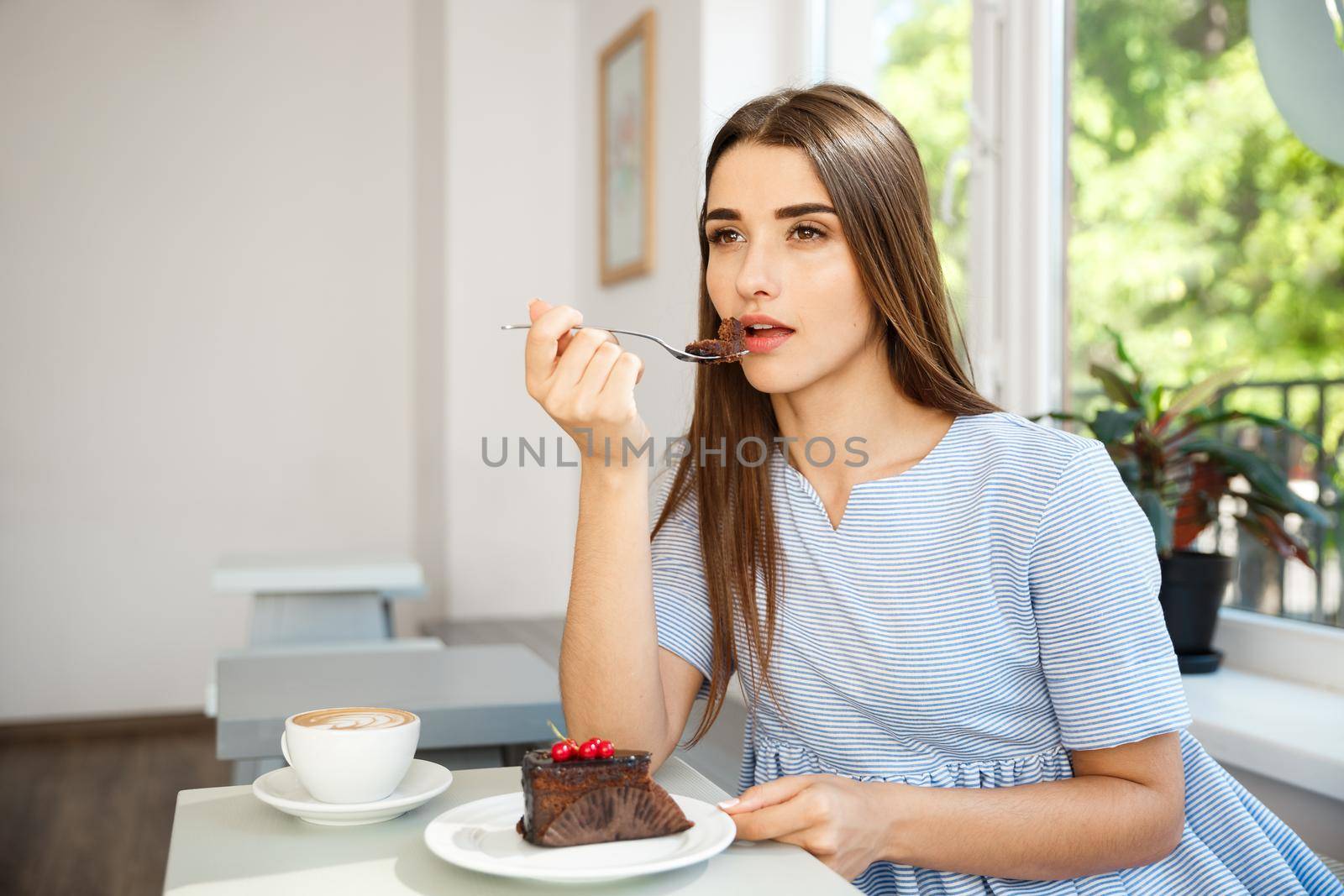 Young attractive Caucasian lady enjoy eating chocolate cake with hot coffee in modern coffee shop at noon. by Benzoix