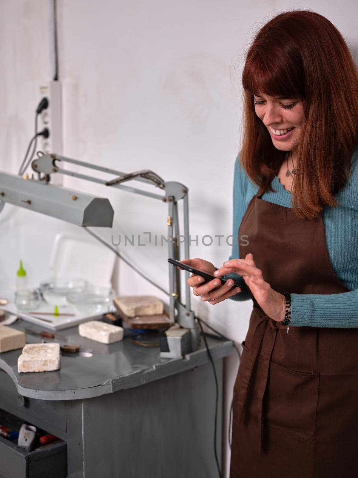 A female worker with apron standing next to her workbench in her jewelry artisan workshop using her phone by WesternExoticStockers