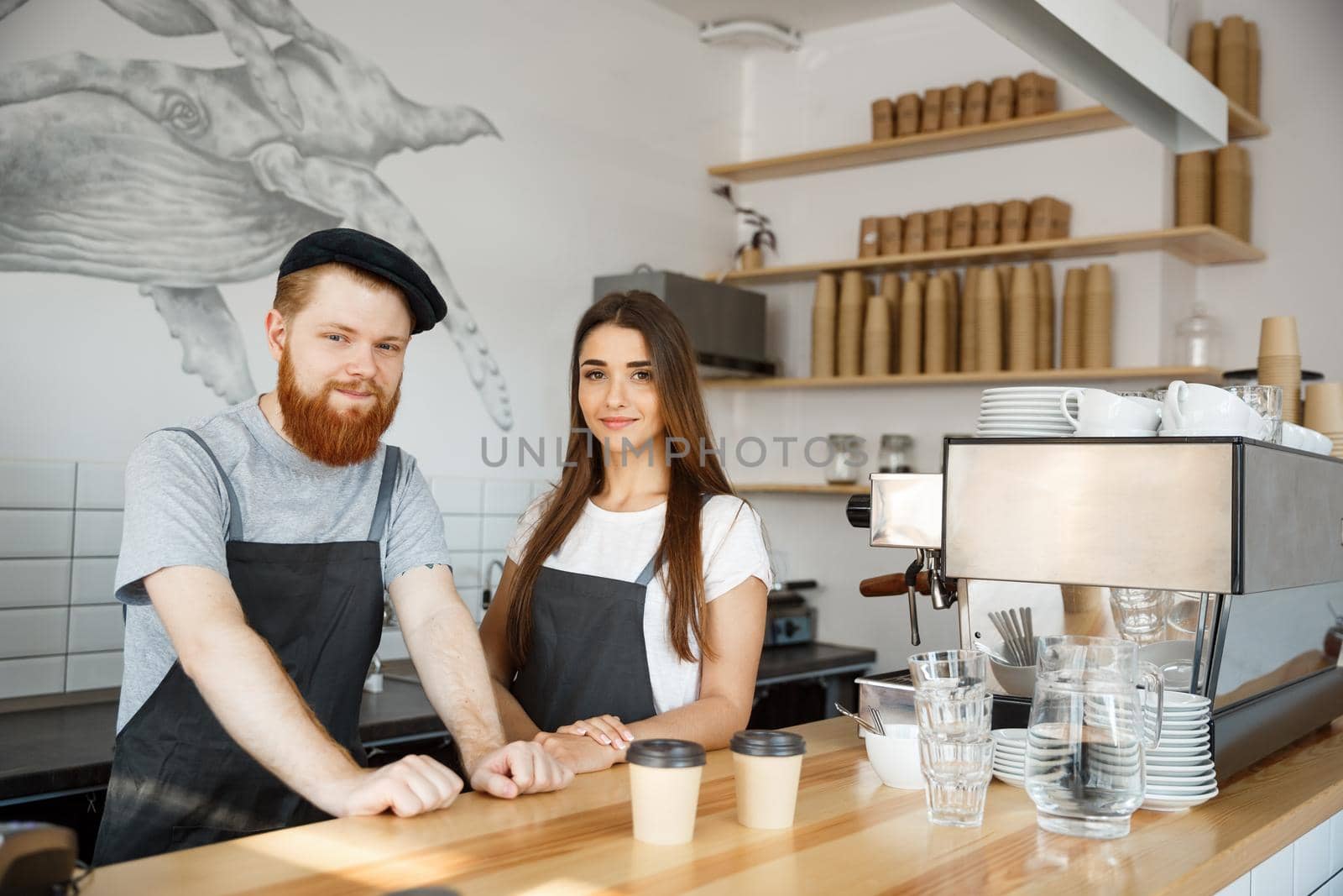 Coffee Business Concept - Positive young bearded man and beautiful attractive lady barista couple in apron looking at camera while standing at bar Couter ready to give Coffee Service at the modern coffee shop