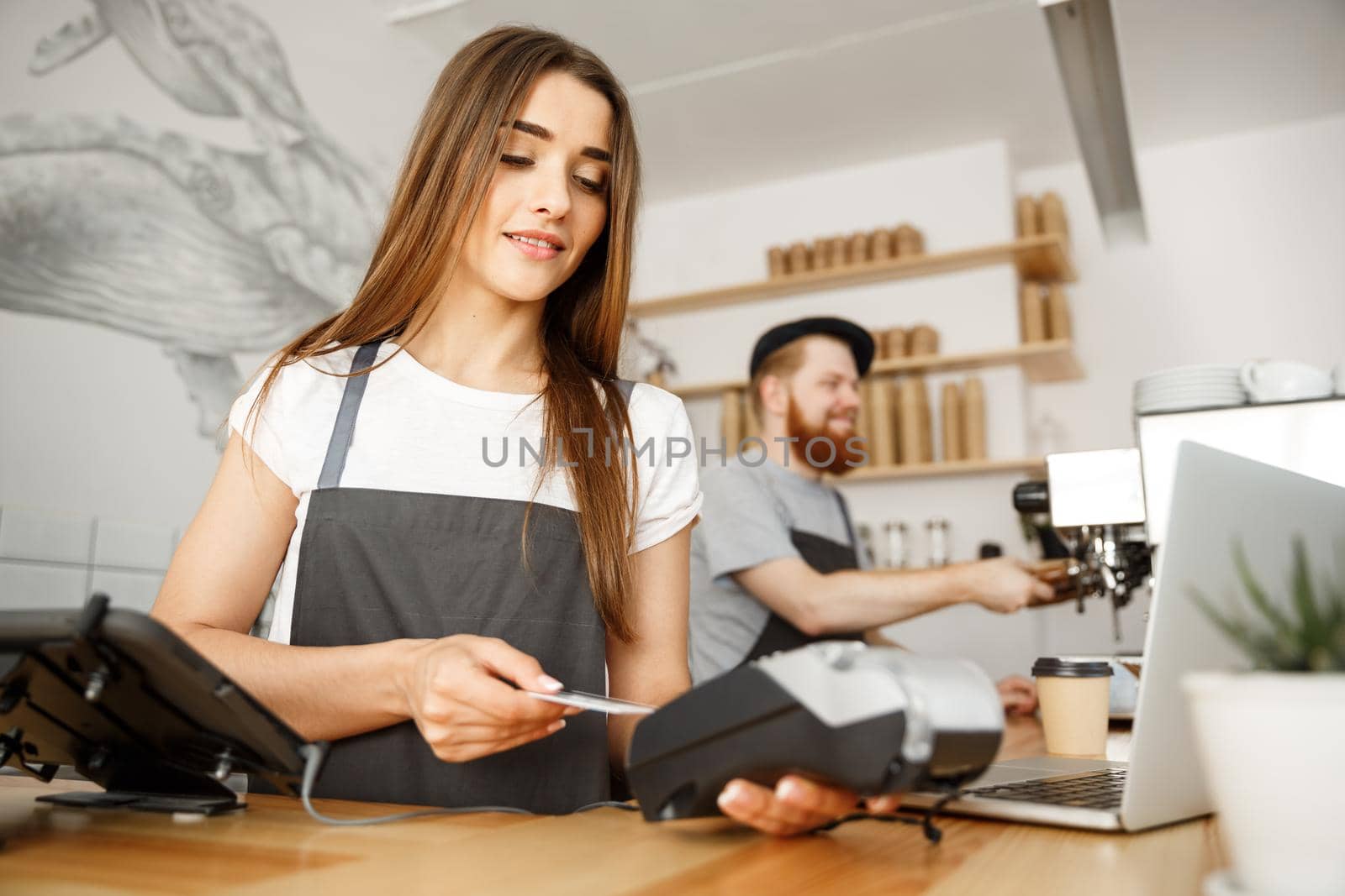 Coffee Business Concept - Beautiful female barista giving payment service for customer with credit card and smiling while working at the bar counter in modern coffee shop. by Benzoix