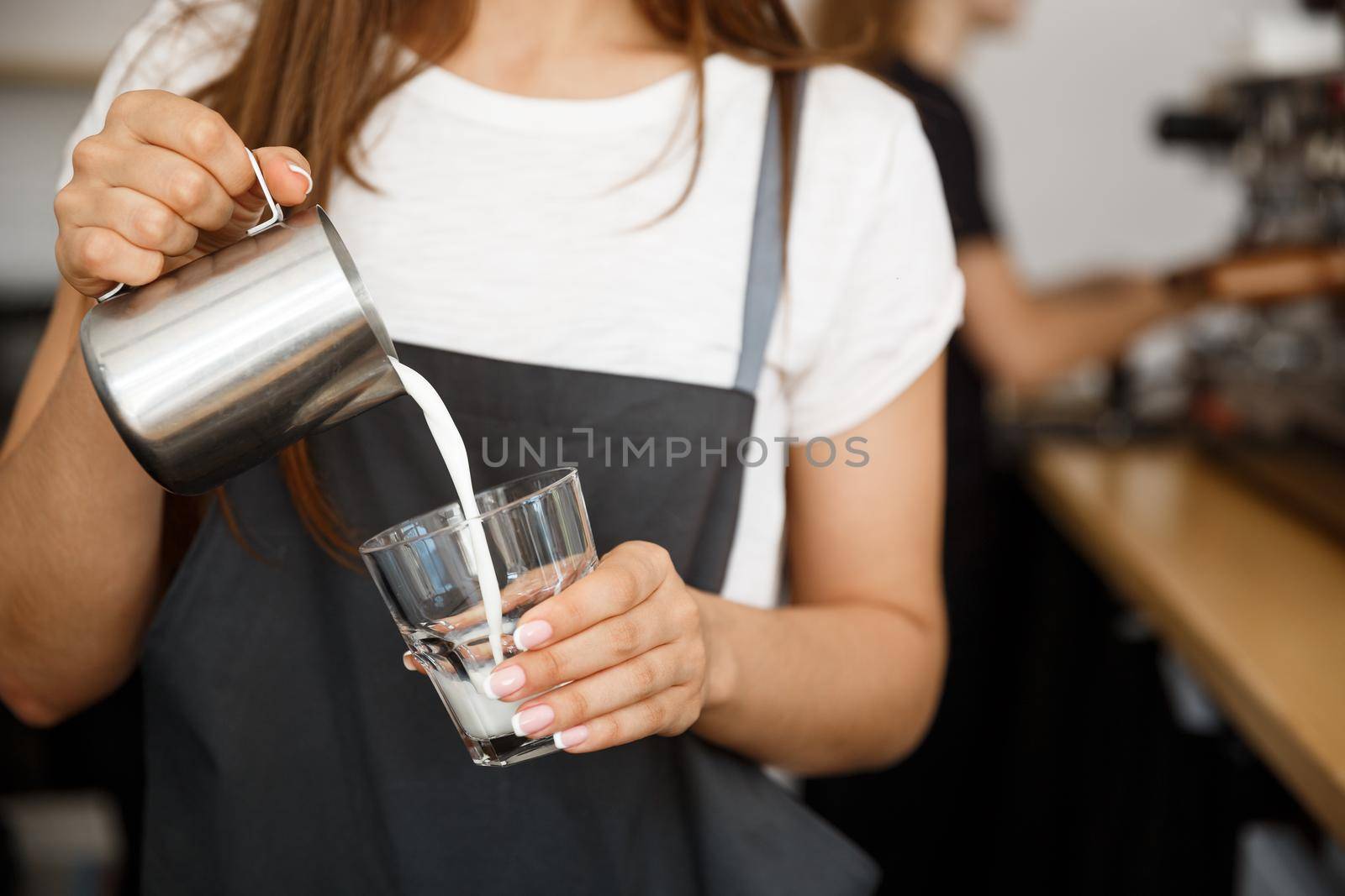 Coffee Business Concept - close-up lady barista in apron preparing and pouring milk in glass cup while standing at cafe.