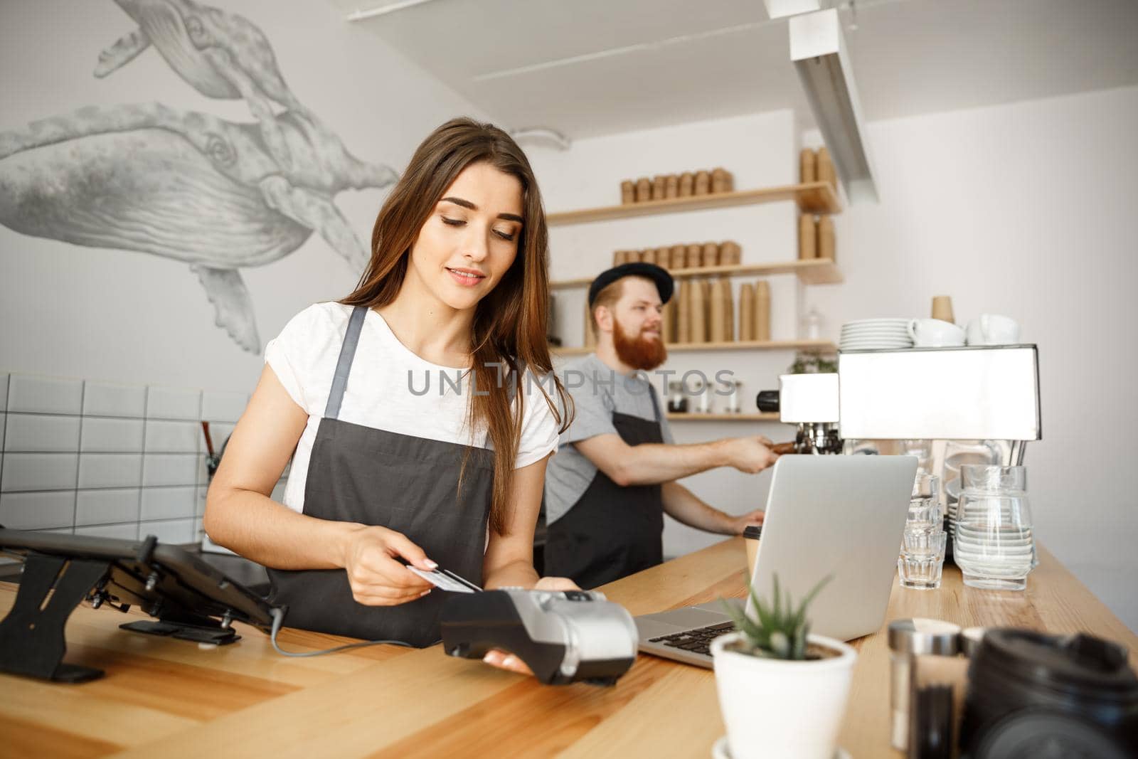 Coffee Business Concept - Beautiful female barista giving payment service for customer with credit card and smiling while working at the bar counter in modern coffee shop. by Benzoix