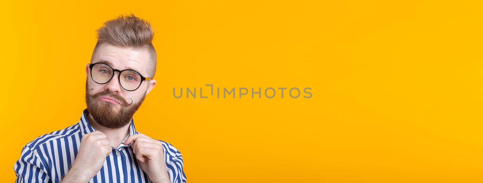 Cute young hipster student guy with a mustache and a beard with glasses straightens his shirt while posing on a yellow background banner with copy space and place for advertising. The concept of self-confidence. by Satura86