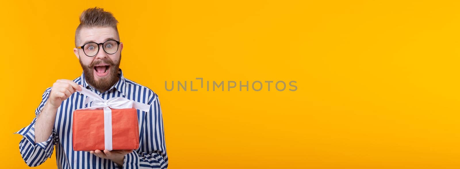 Surprised young hipster man with mustache and beard in surprise unpacks a red box with gifts on a yellow background banner with copy space, close up. The concept of gifts and surprises for the holiday by Satura86