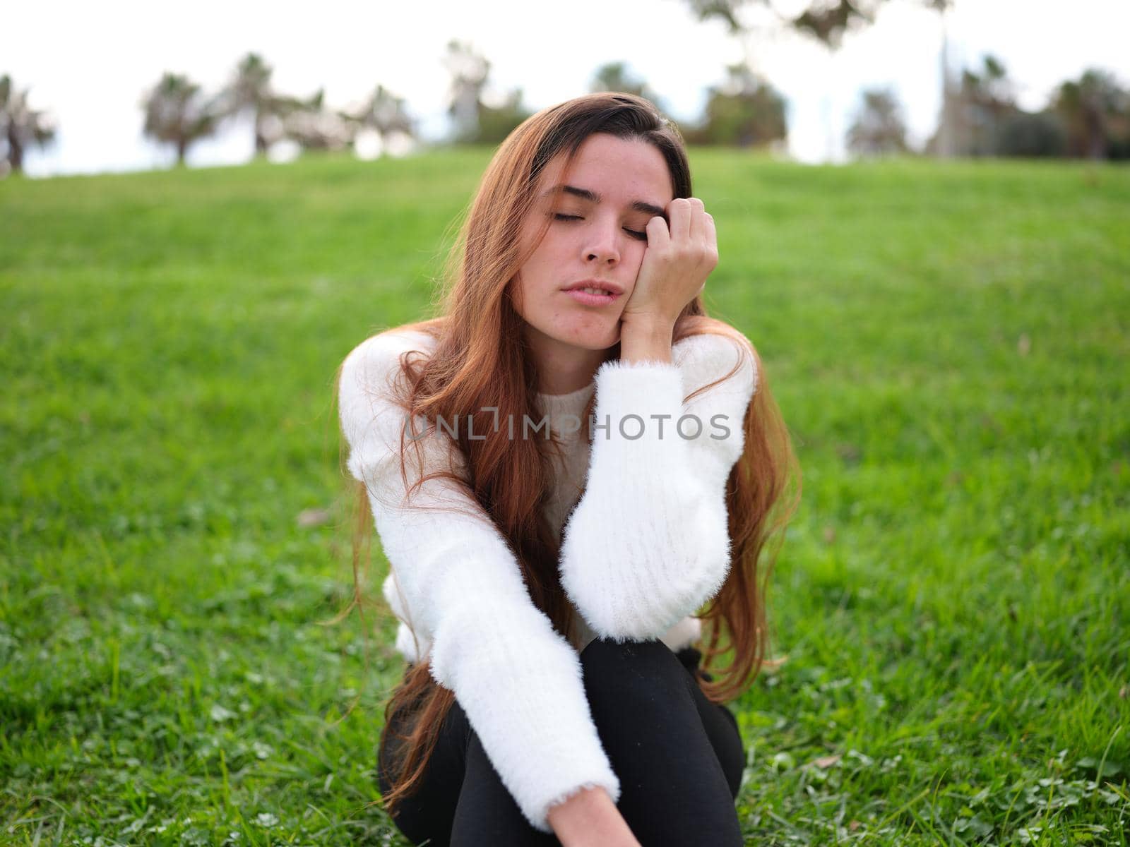 Front view of a young woman in the park sitting on the grass with one hand on her face falling asleep. Concept of emotions.