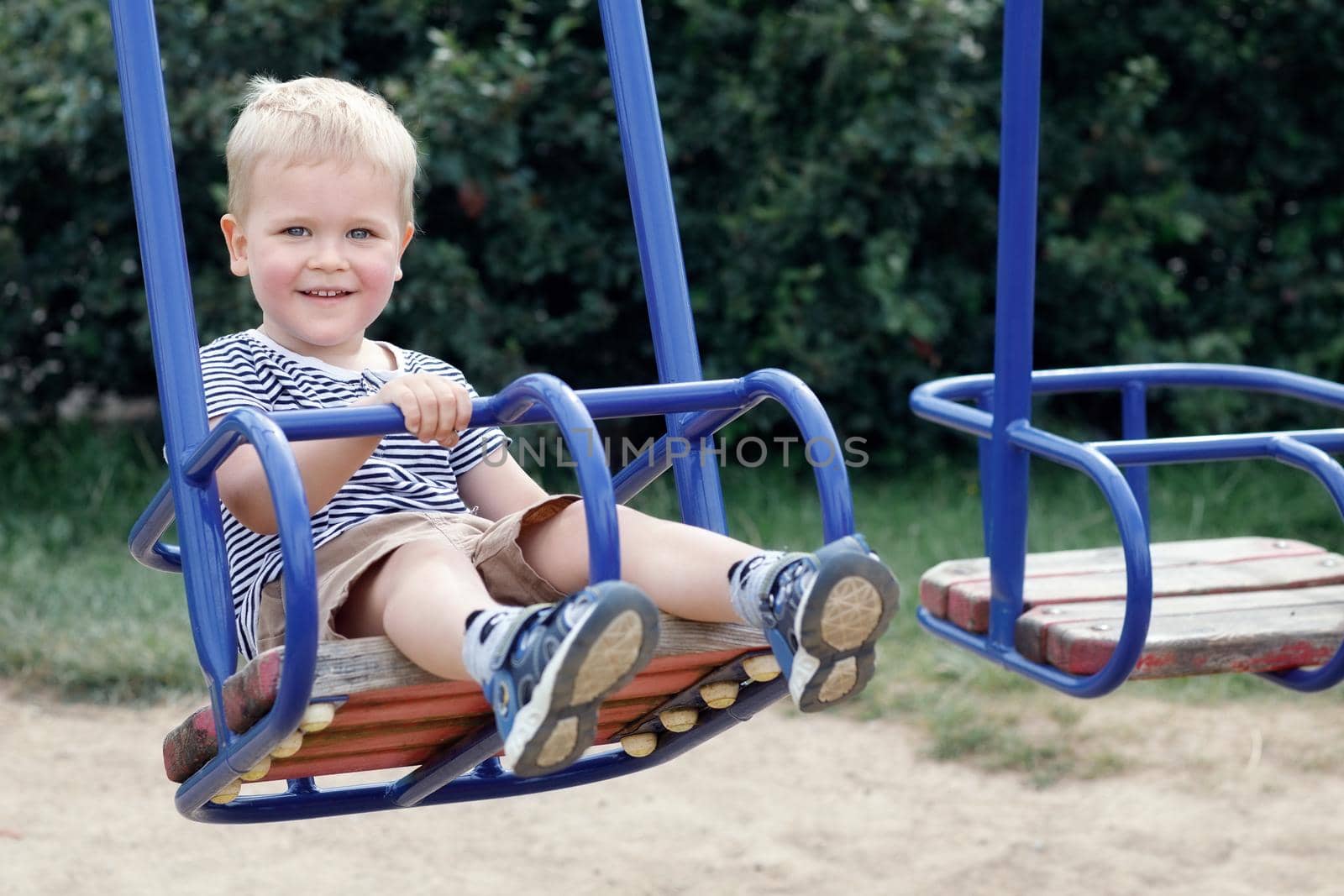 Little boy swinging on the swings of a public playground. Looking at camera and smiling. Happy childhood