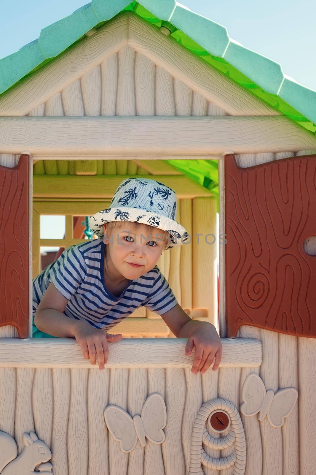 A cheerful little boy in a hat, inside a toy little house, he looks out the window with the shutters straight into the camera