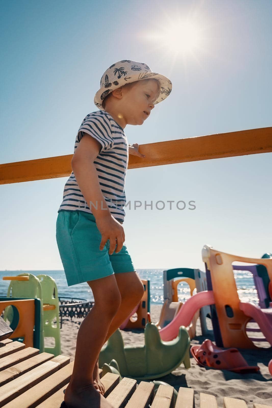 A cute boy in a hat walks on a wooden bridge to the beach. A playground, sea and sun rays in the background