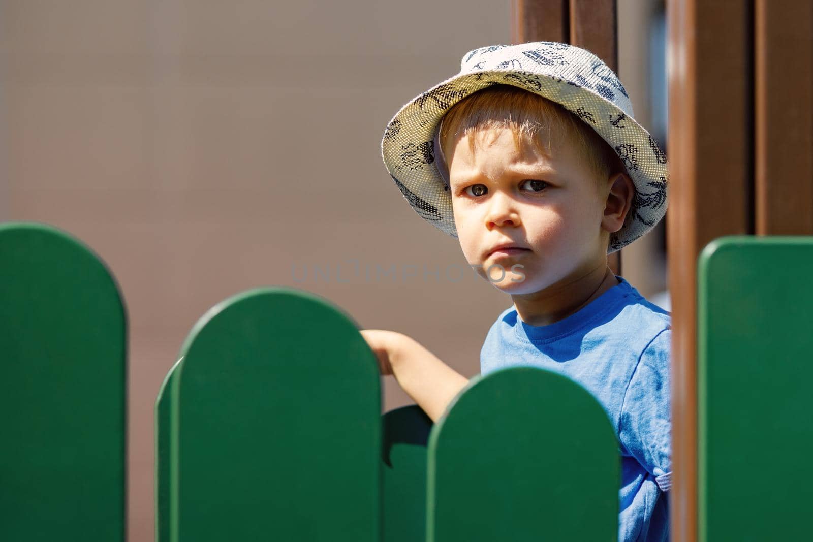Little caucasian preschool boy with hat playing in playground in city public park near green fence
