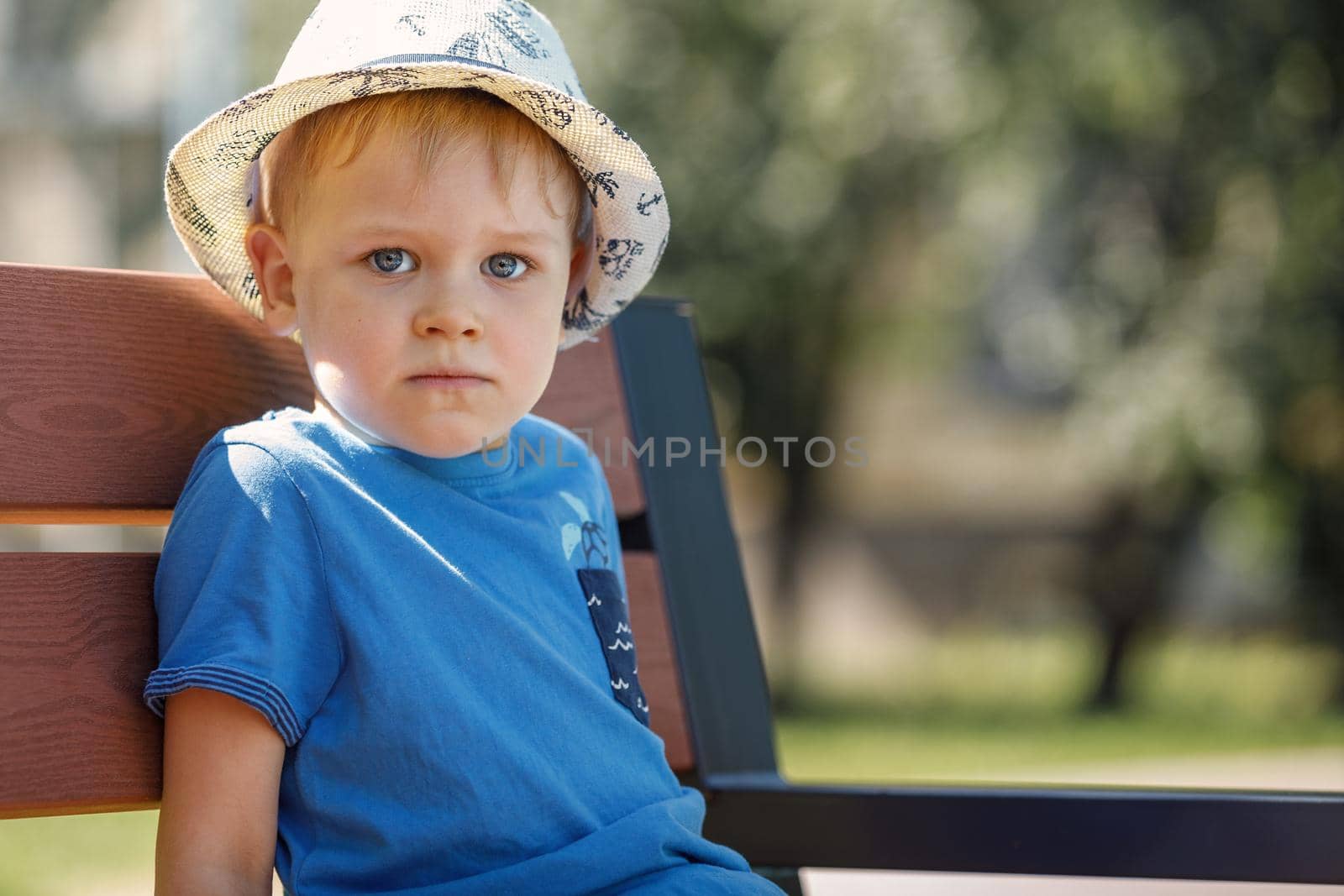 Portrait of a cute boy with a white hat and a blue shirt. A child sits on a nat bench in the park and looks straight at the camera